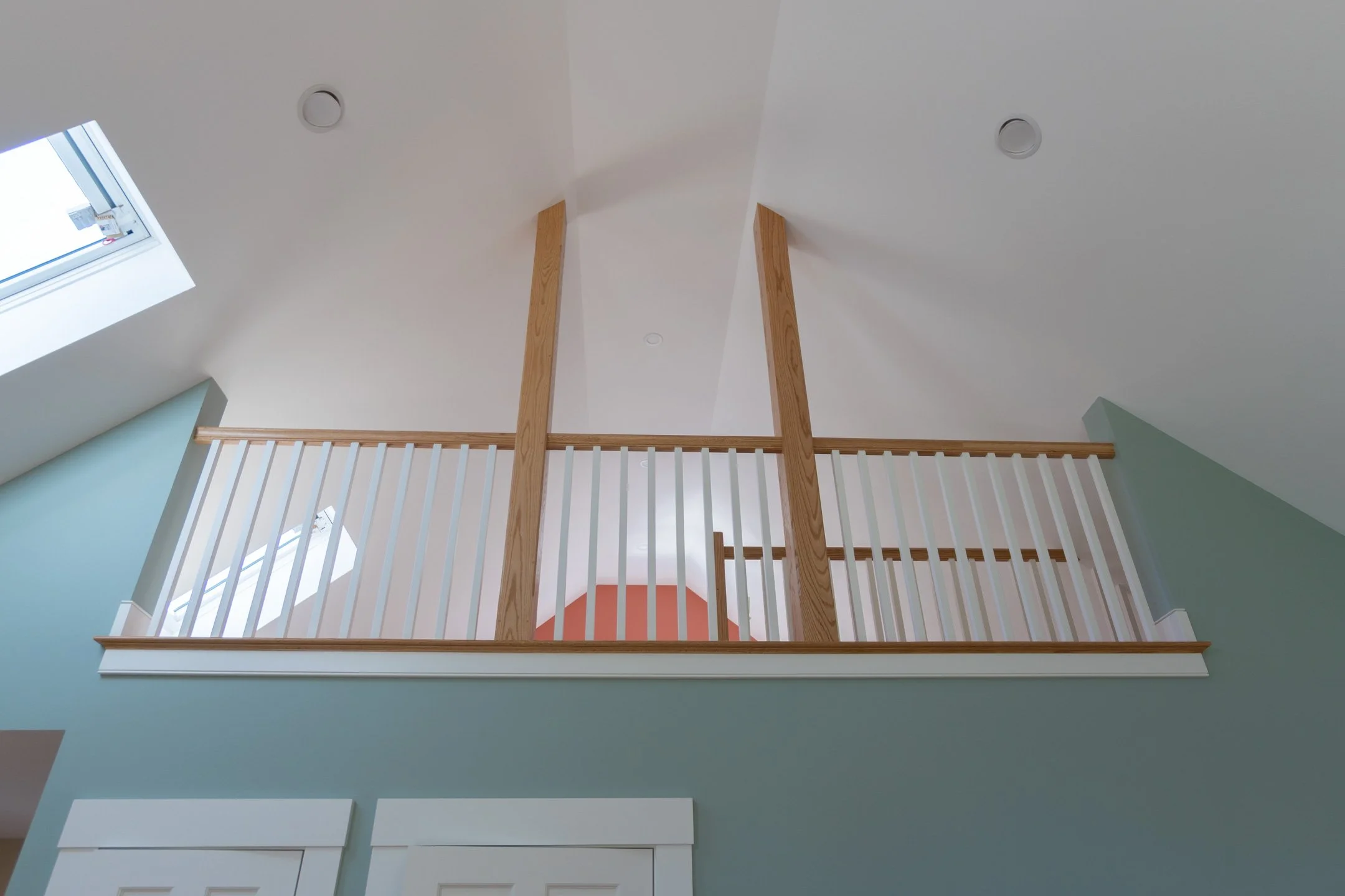 Interior view of a modern home showcasing a wooden balcony railing and support beams, with a sloped ceiling featuring skylights and recessed lighting.