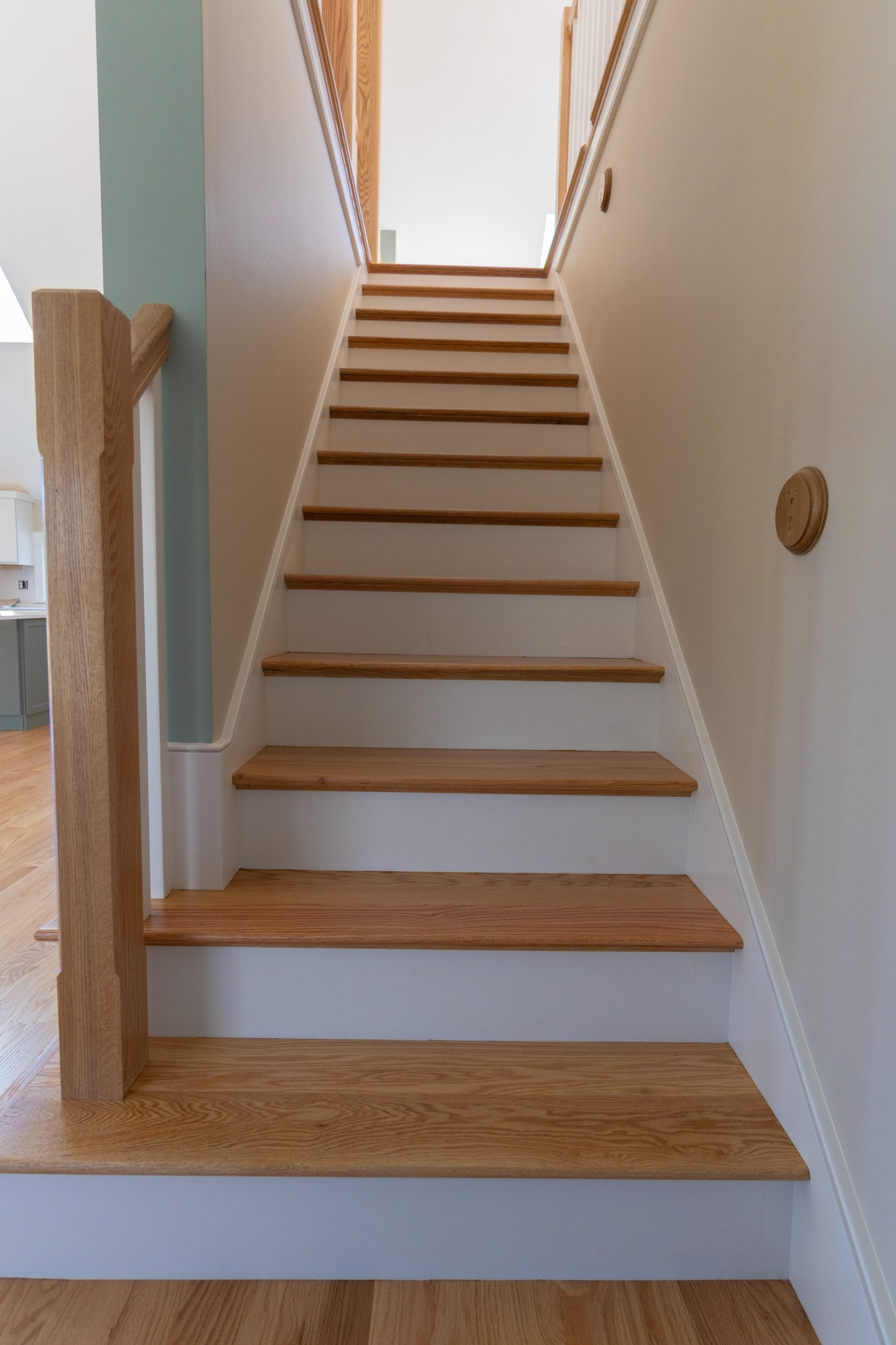 A staircase with natural wood steps and a wooden handrail on the left side, leading up to an upper floor with a light-colored wall and a wooden frame opening at the top.