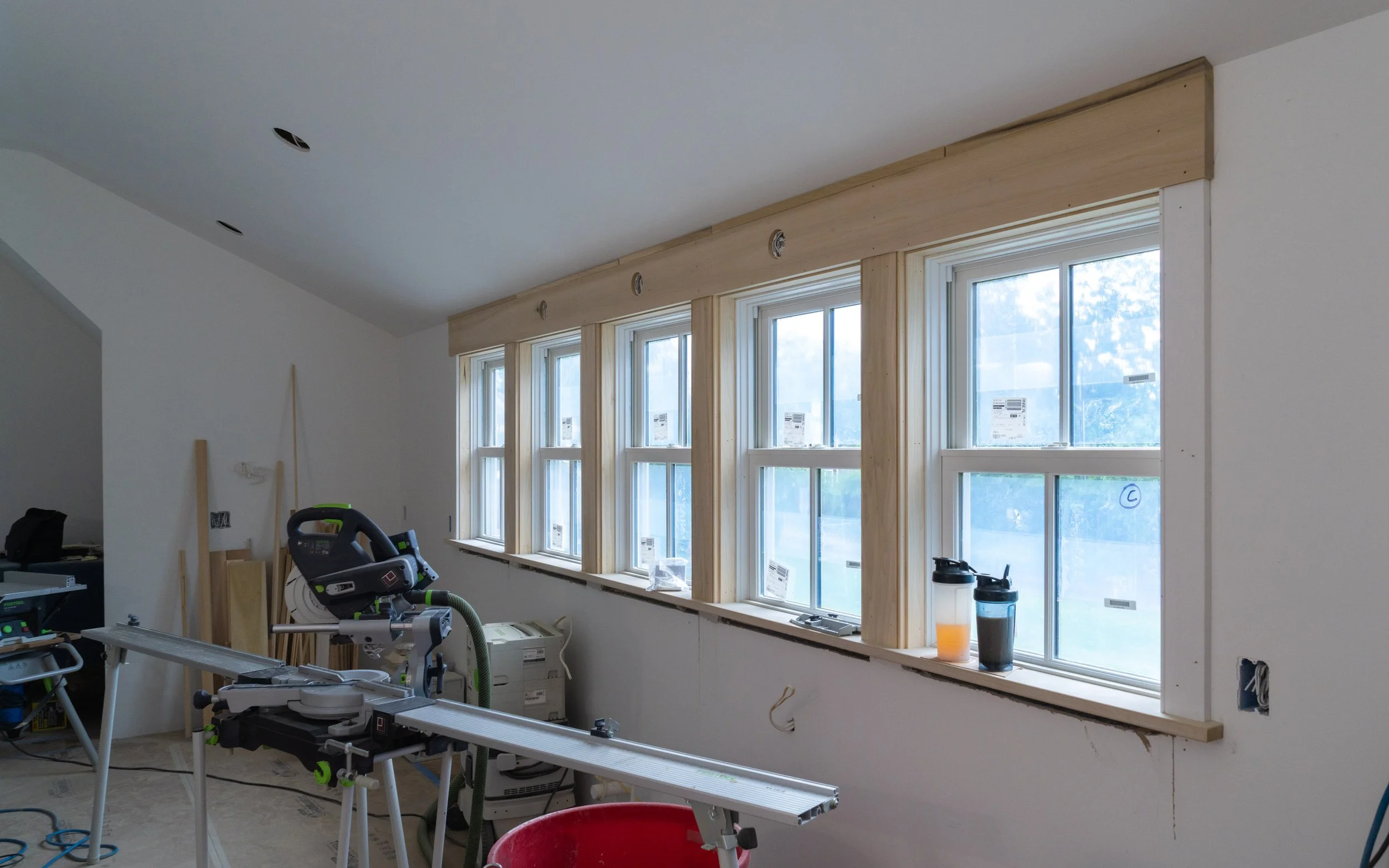 Room under construction with large windows, tools, and equipment, including a saw, drill, and buckets, with unfinished walls and ceiling.