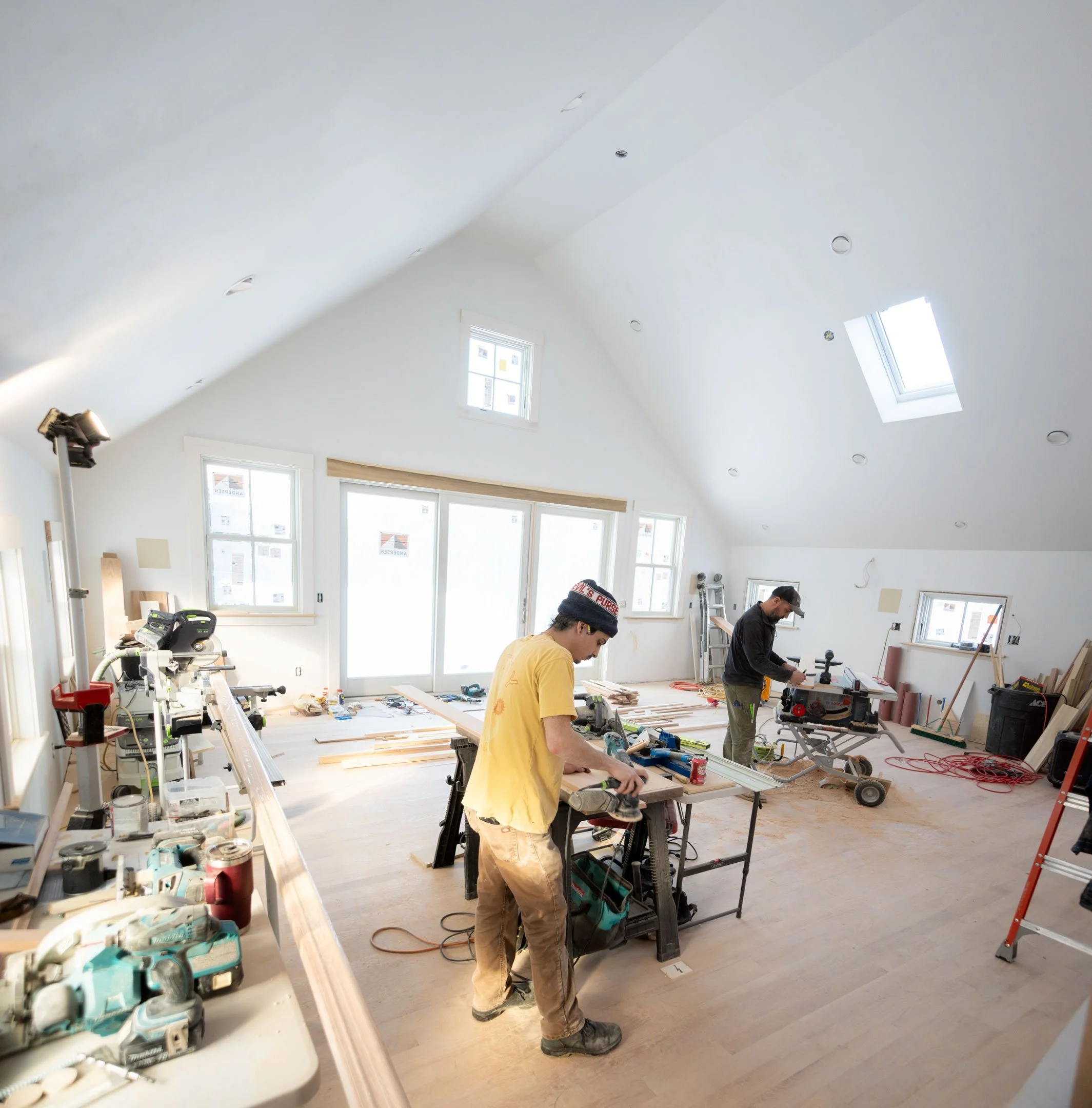 Interior of a room under construction with two workers working. Tools and equipment are spread across a workbench and the floor, with windows letting in natural light.