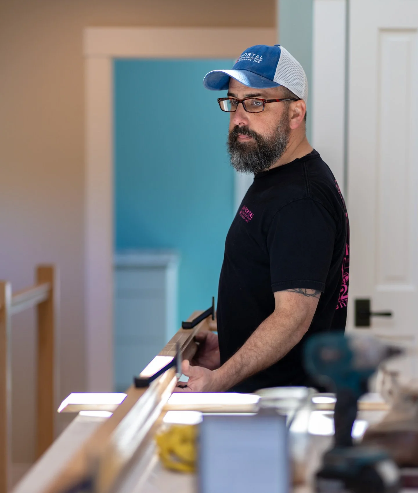 Man with glasses and a beard, wearing a blue and white cap and a black t-shirt, appears to be working in a workshop or store, holding an object and looking to his right.