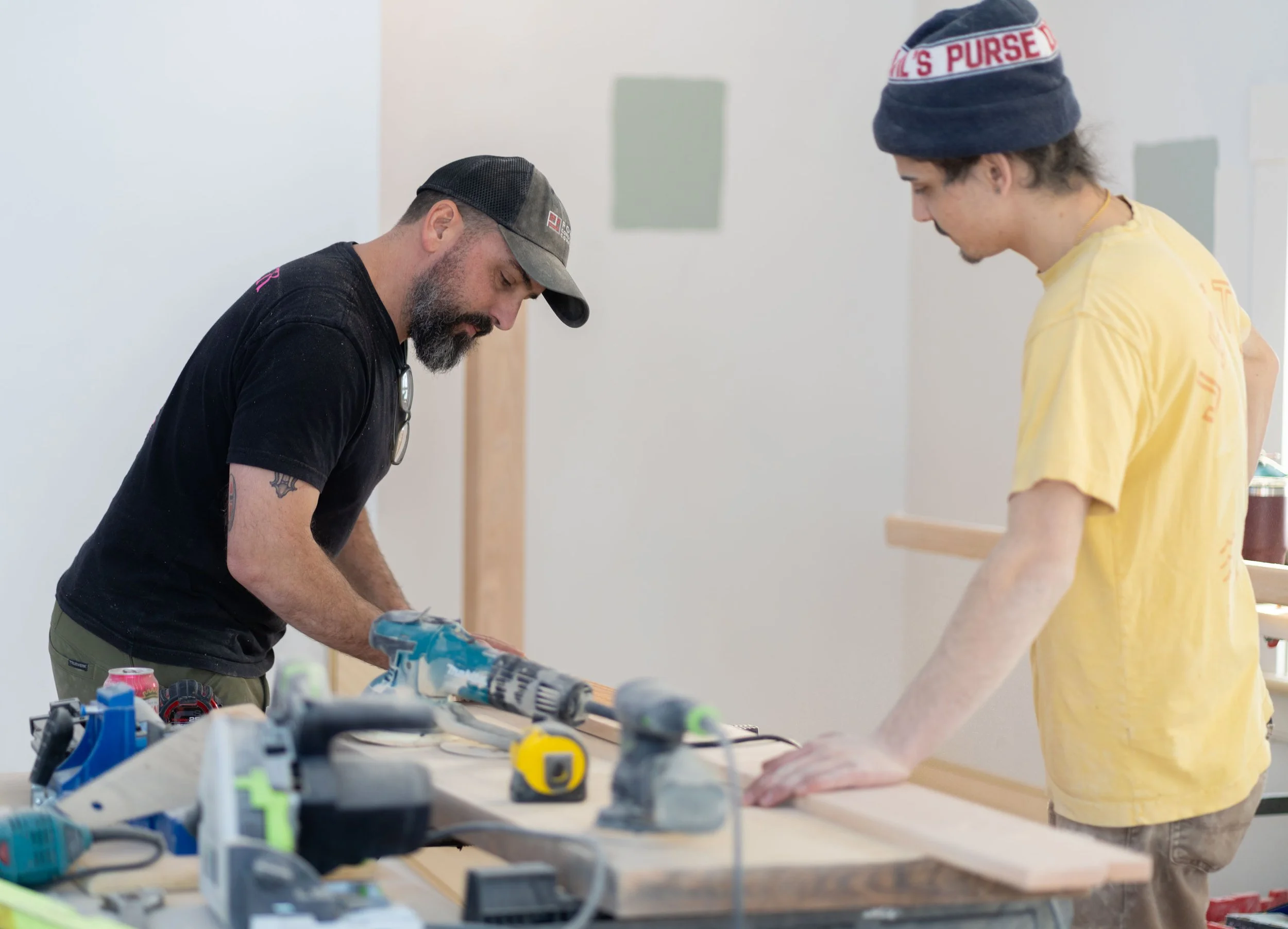 Two men working on a project at a table with tools and other items. One man is wearing a black shirt and cap, the other is in a yellow shirt and beanie.