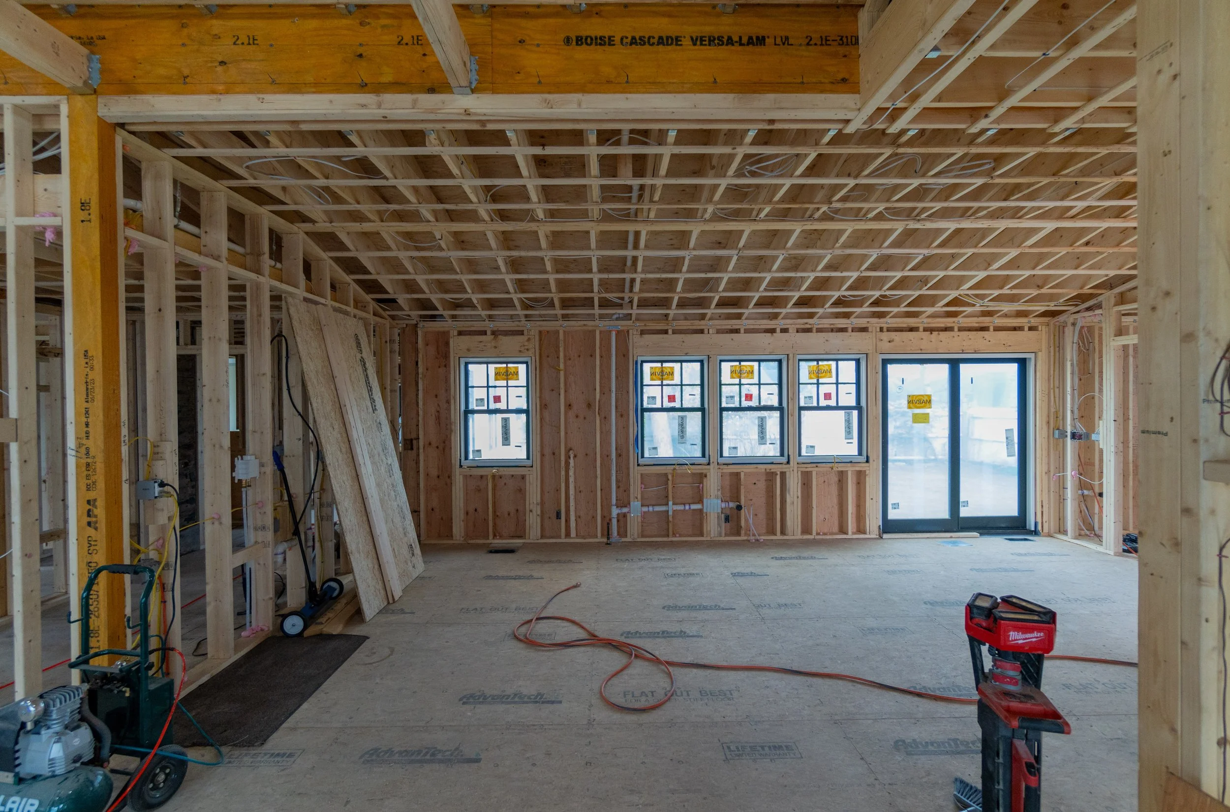 Interior of a house under construction showing wooden framing, exposed ceiling joists, and windows. Construction tools and materials are visible on the floor.