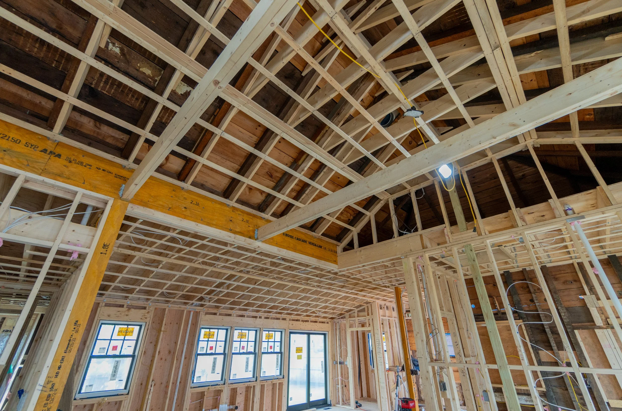 Interior view of a house under construction, showing wooden framing, window openings, and overhead wiring.