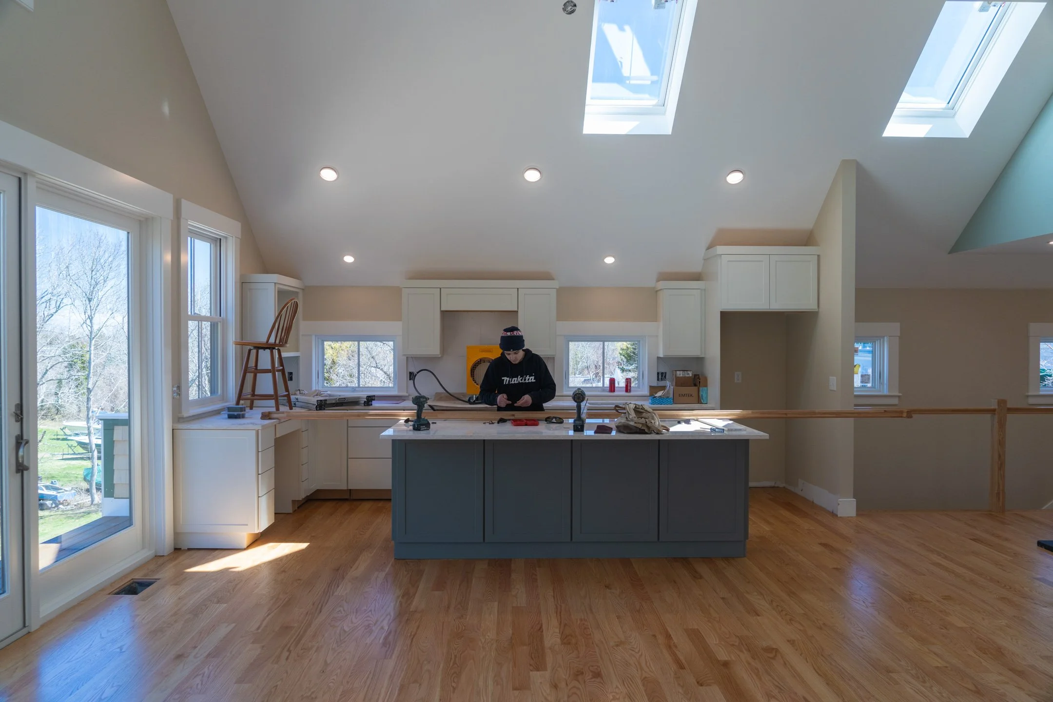 Open kitchen with white cabinets, a blue island, hardwood floors, large windows, and skylights, with a person working at the counter.