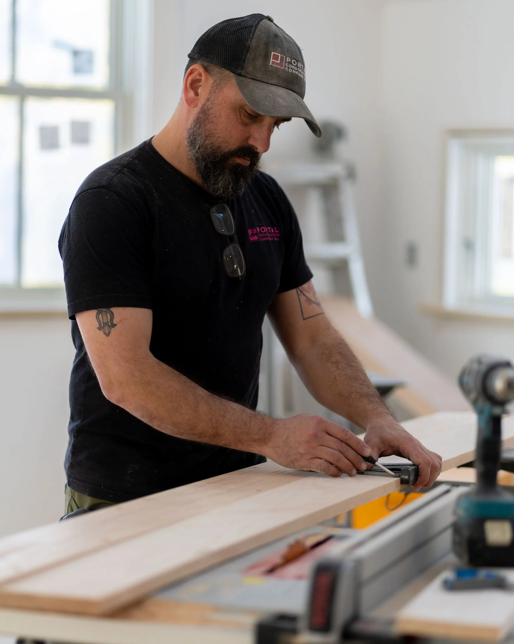 A man with a beard and tattoos wearing a gray and black baseball cap and a black t-shirt working on a piece of wood at a workshop.