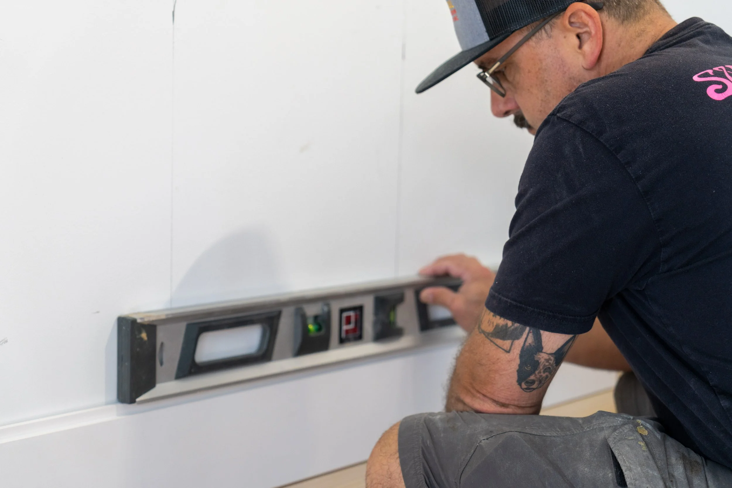 A man with tattoos on his arm, wearing a dark T-shirt and a baseball cap, is installing or repairing an electrical outlet or switch on a white wall, with a level tool placed underneath for alignment.