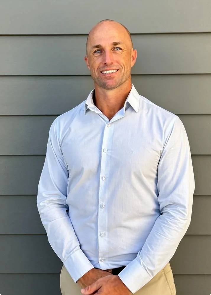 A smiling man with a shaved head wearing a light blue dress shirt and beige pants standing outdoors in front of a gray horizontal siding wall.