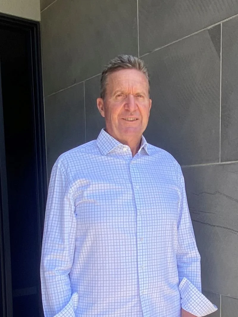 A middle-aged man with short, light brown hair stands outside near a modern gray stone wall, wearing a white and blue checkered dress shirt.