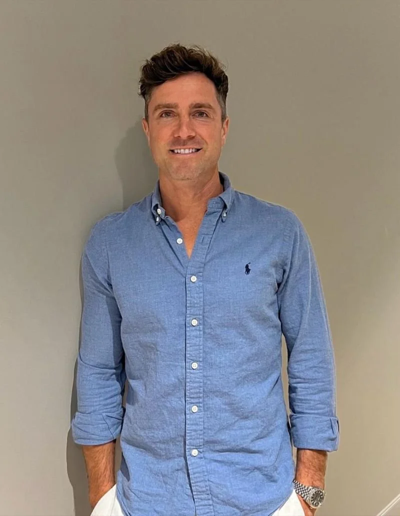 A man with brown hair, wearing a blue button-up shirt with rolled-up sleeves, standing against a plain gray background.