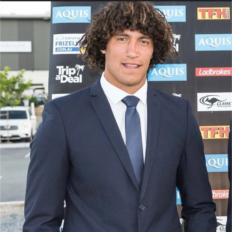 A man with curly brown hair wearing a dark blue suit, white shirt, and dark blue tie, standing outdoors in front of a backdrop with various logos and sponsors.