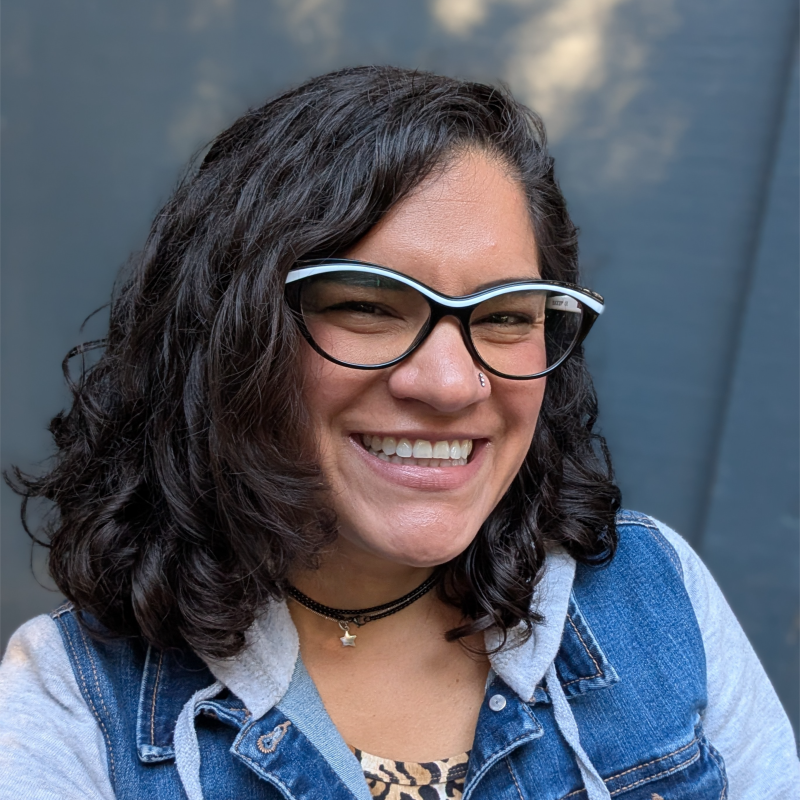 A woman with long brown wavy hair smiling, standing outdoors against a beige wall and a window, wearing a black top, patterned gray blazer, and small hoop earrings.