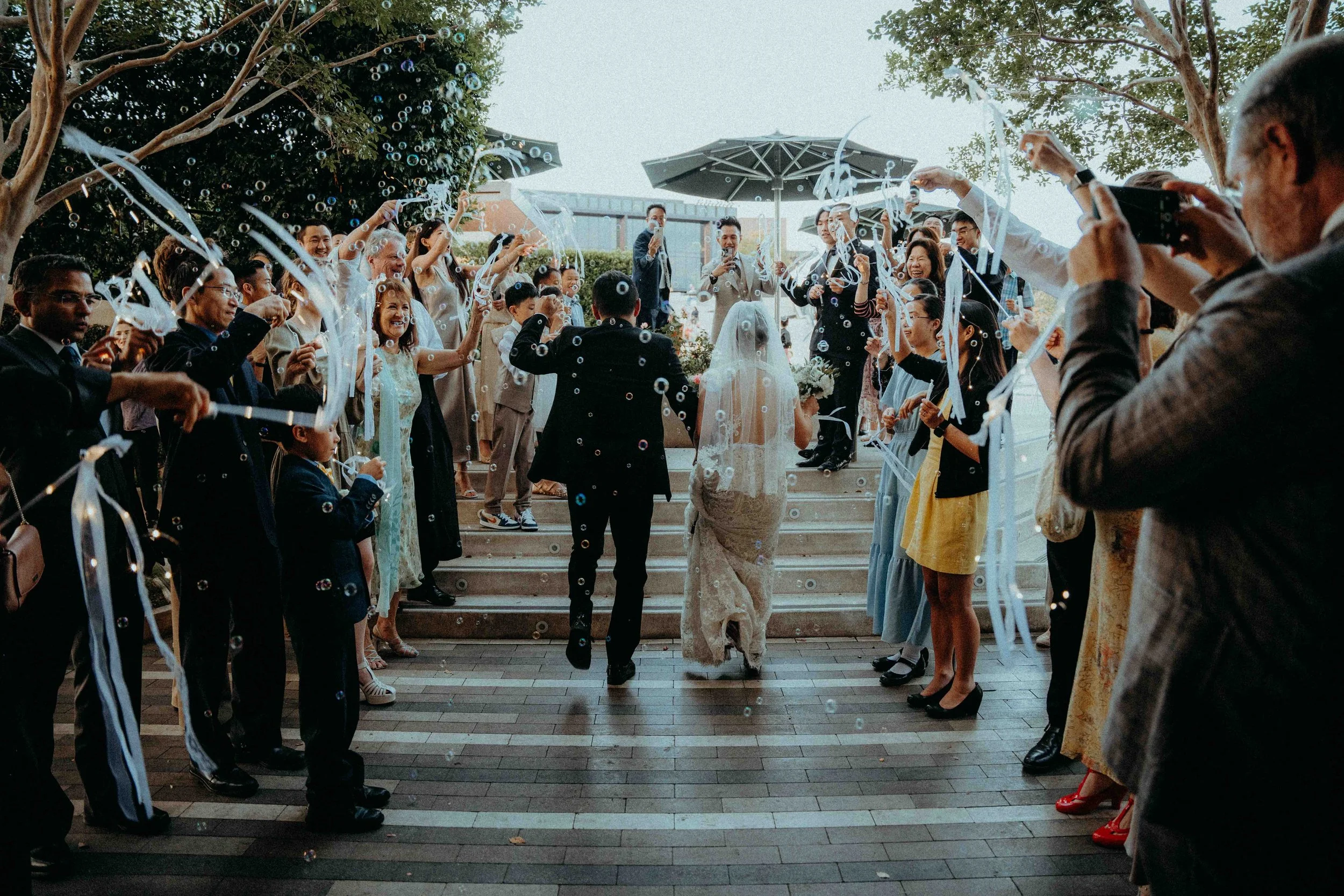 Bride and groom walking away from a wedding celebration under an arch, surrounded by guests holding ribbons and bubbles, standing on steps outdoors.