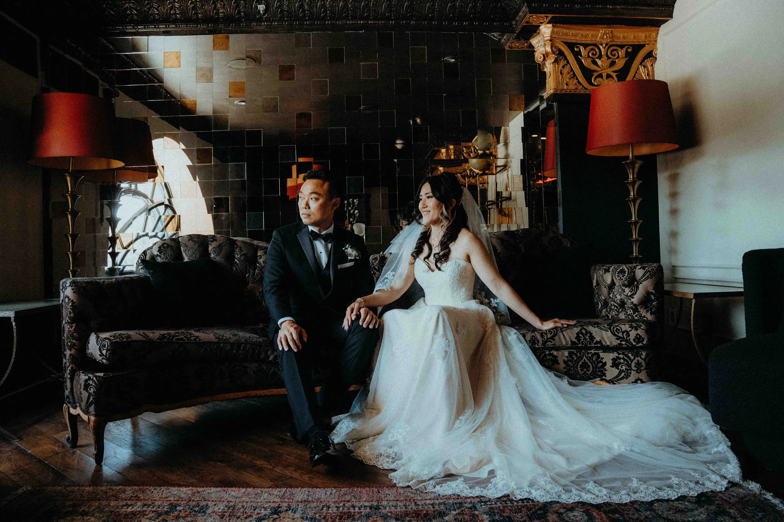 A bride in a white wedding gown and a groom in a black tuxedo seated on a vintage sofa in an elegant room with dark wood flooring, gold accents, and large lamps with red shades.