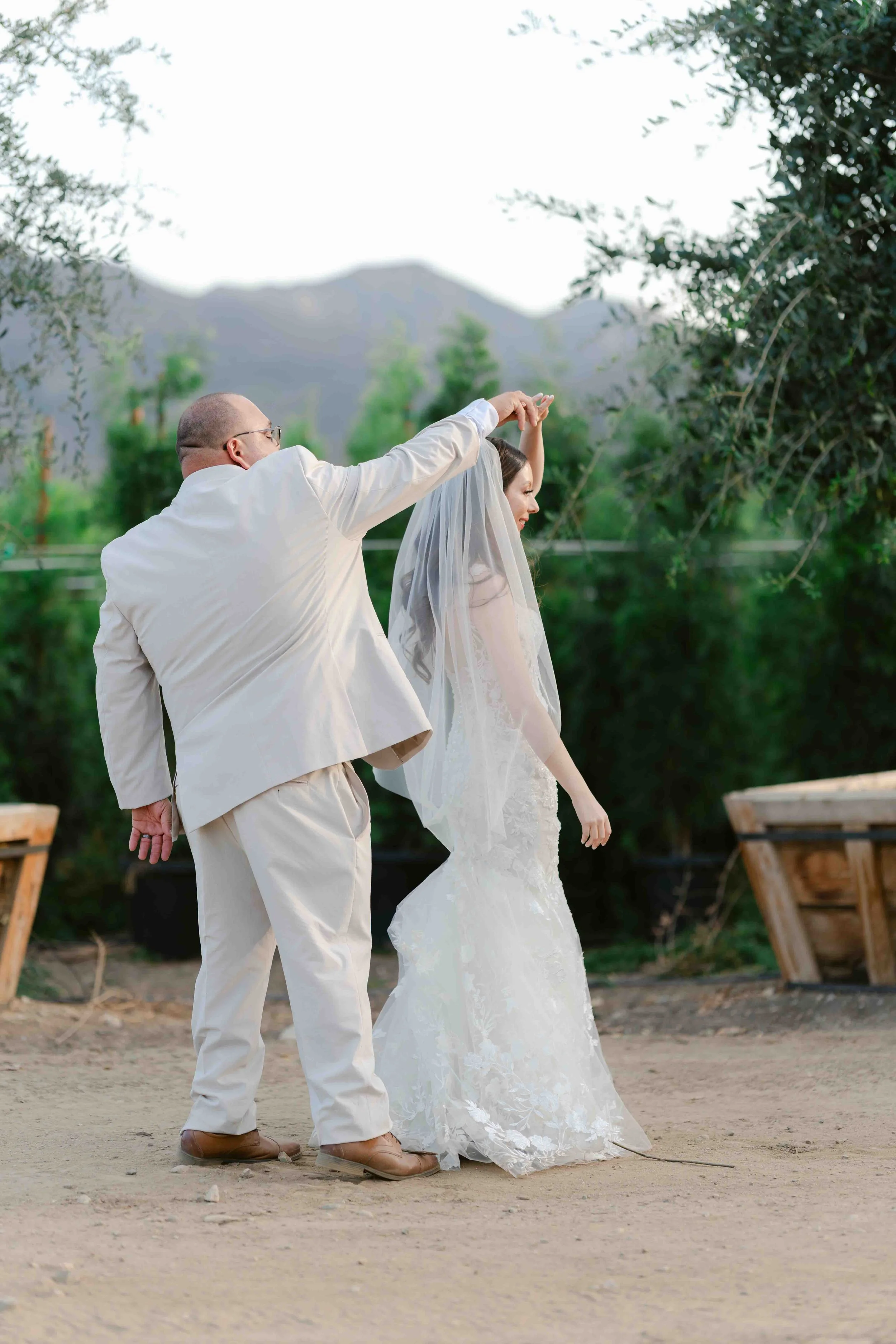 A man and woman in wedding attire dancing outdoors, with the man spinning the bride, set against mountains and greenery.
