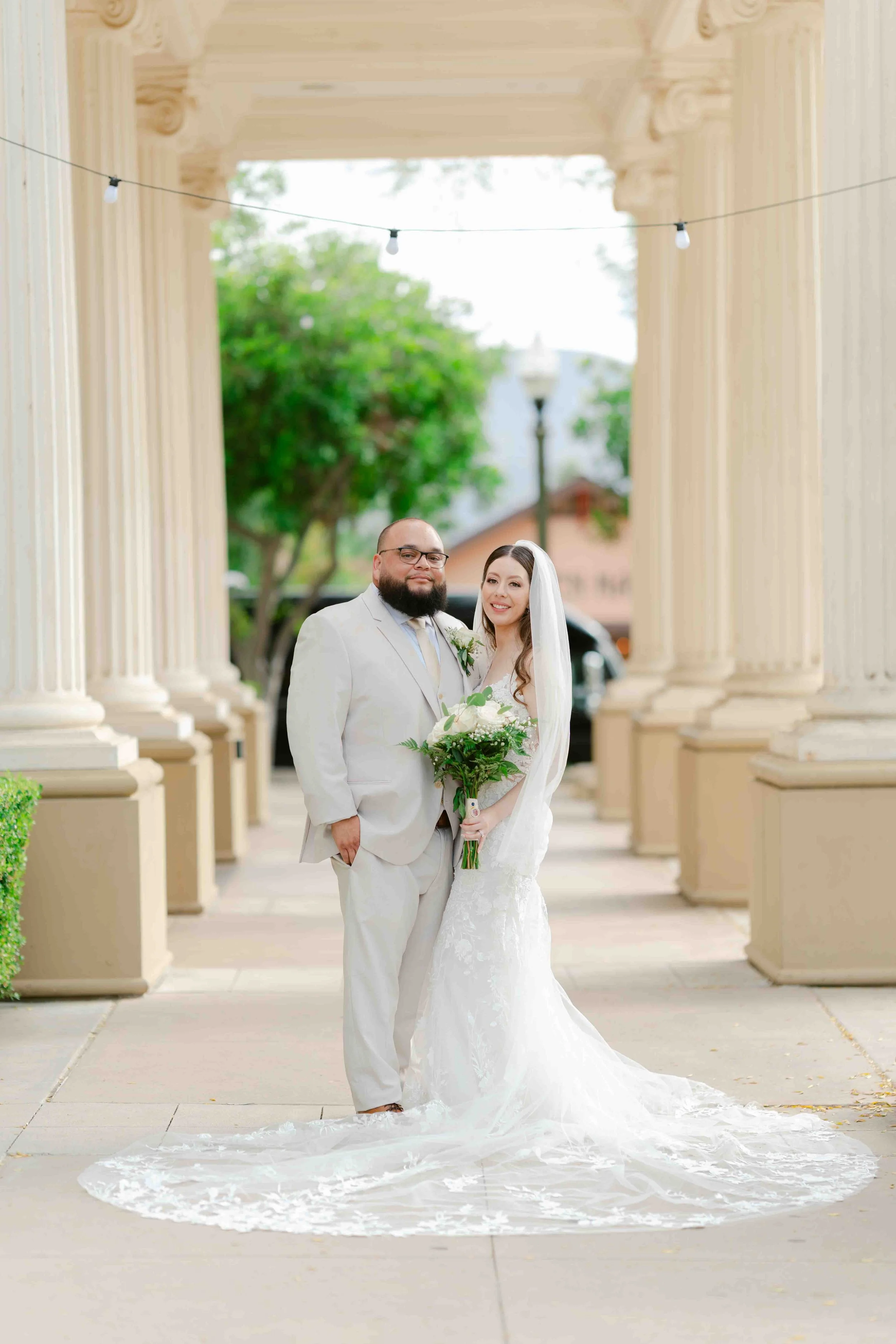 A bride and groom standing under an archway, both dressed in wedding attire, with the bride holding a bouquet of white flowers.