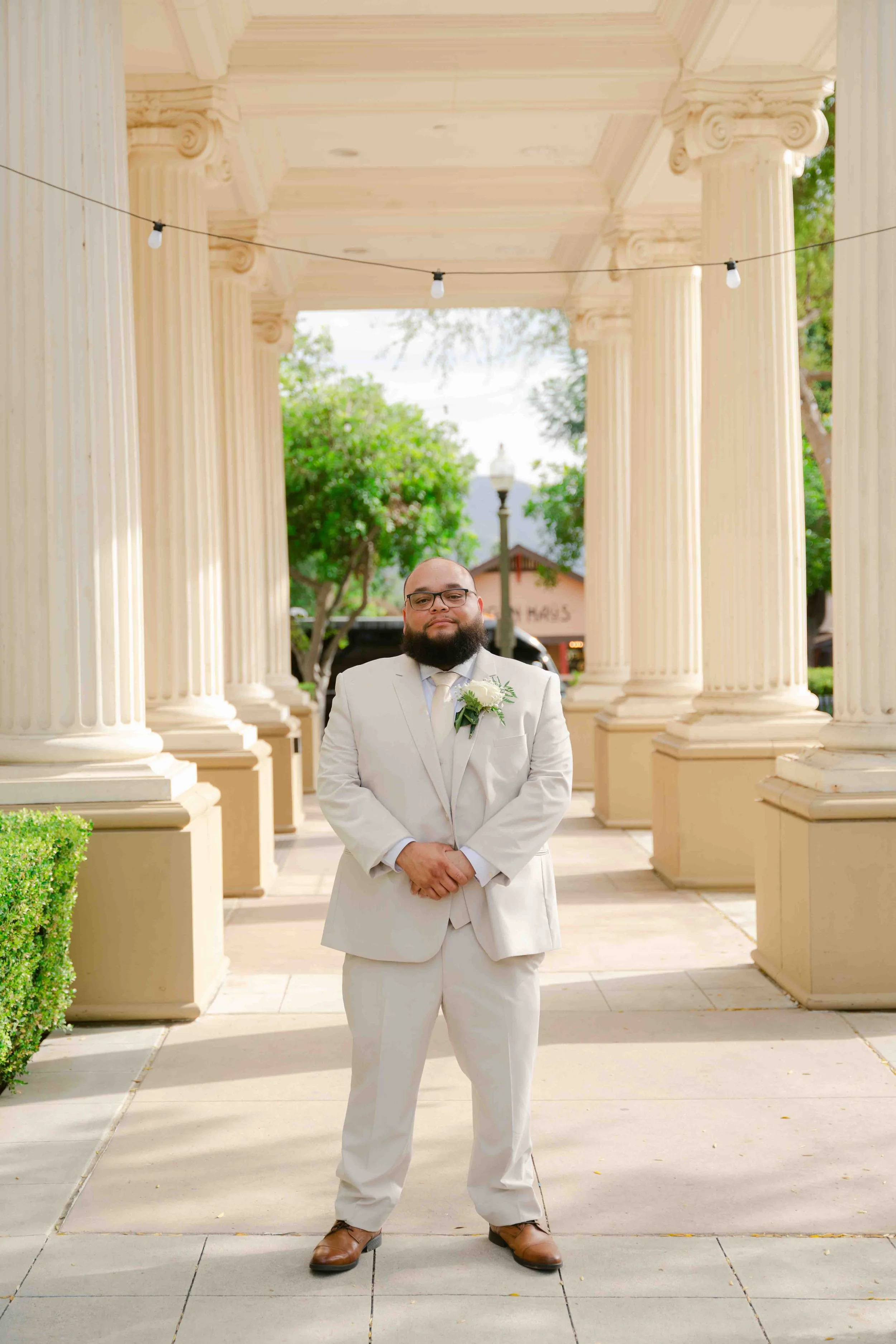 A man in a light-colored suit and glasses standing outdoors in front of large classical columns with greenery in the background.