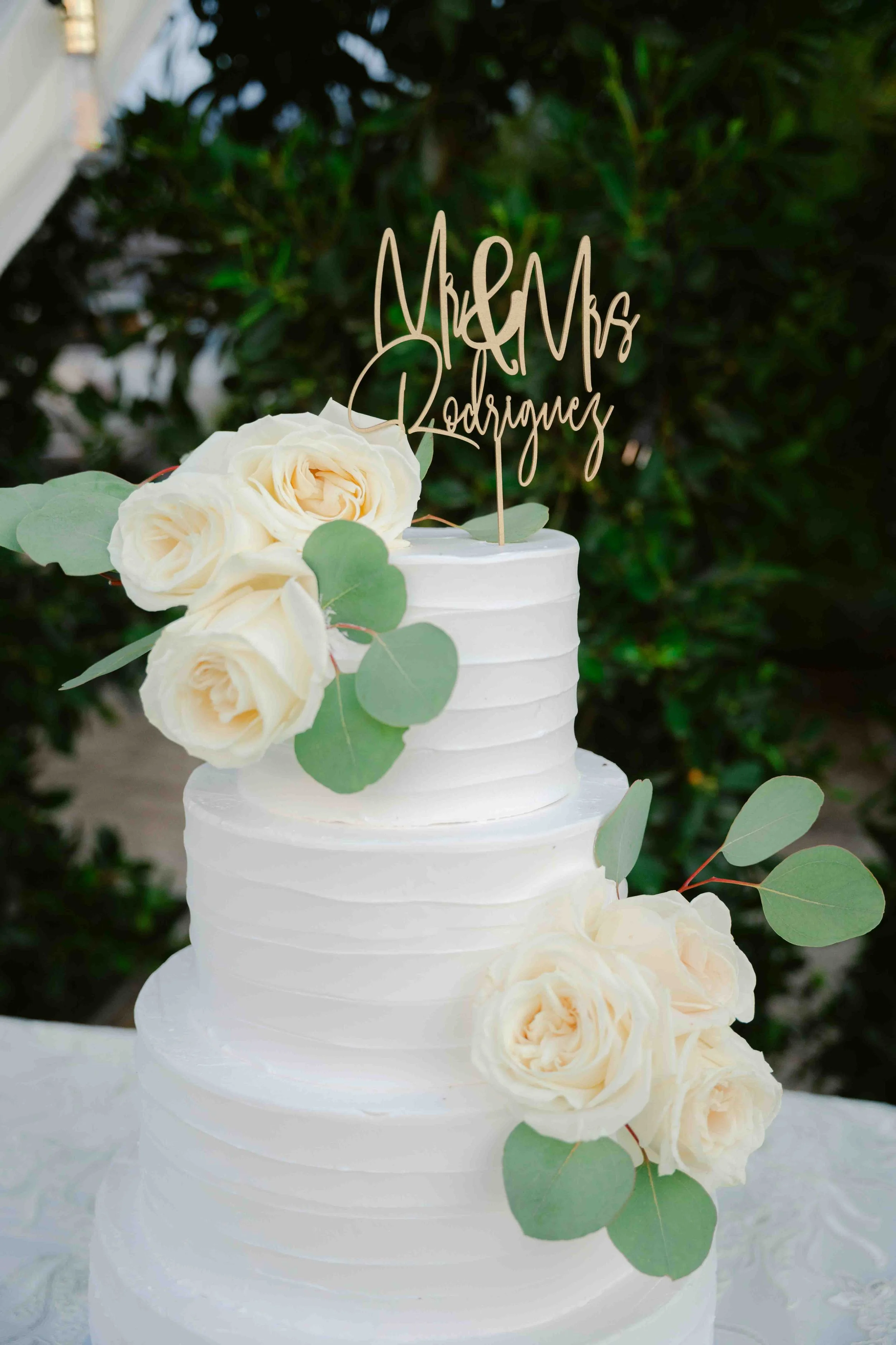 A three-tiered white wedding cake decorated with white roses and eucalyptus leaves, with a gold-colored cake topper that says 'Mr & Mrs Ladriores'.
