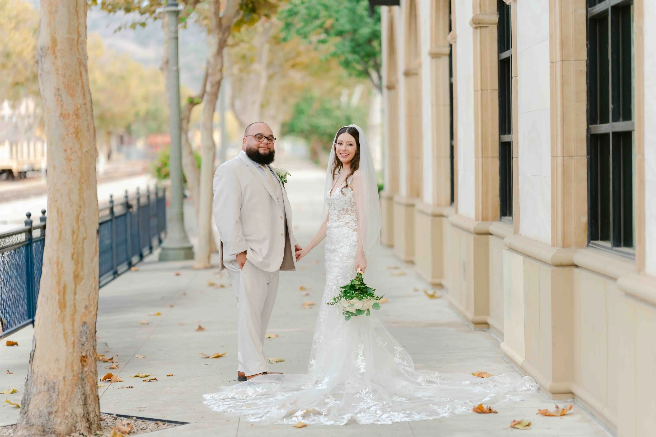 A newlywed couple stands hand in hand on a city sidewalk lined with trees and historic buildings, with the groom wearing a light-colored suit and the bride in a white lace wedding dress holding a bouquet, both smiling at the camera.