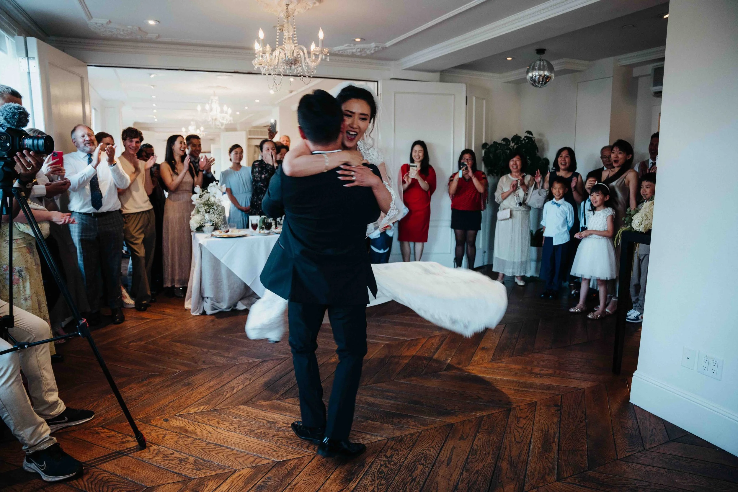 A wedding reception with a bride and groom dancing together, surrounded by family and friends who are clapping and smiling, in a decorated indoor venue.