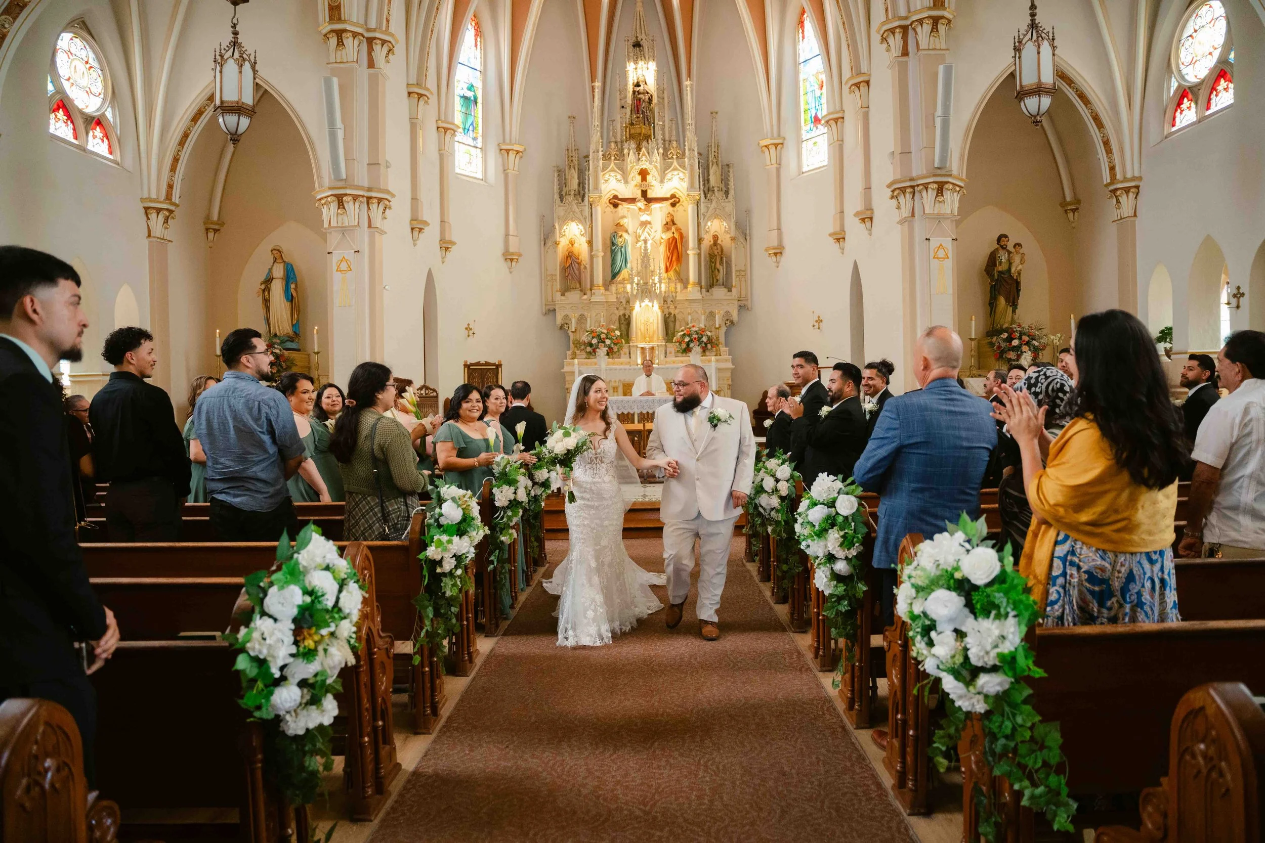 A bride and groom walking down the aisle at their wedding ceremony inside a church, surrounded by guests and decorated with white flowers and greenery.