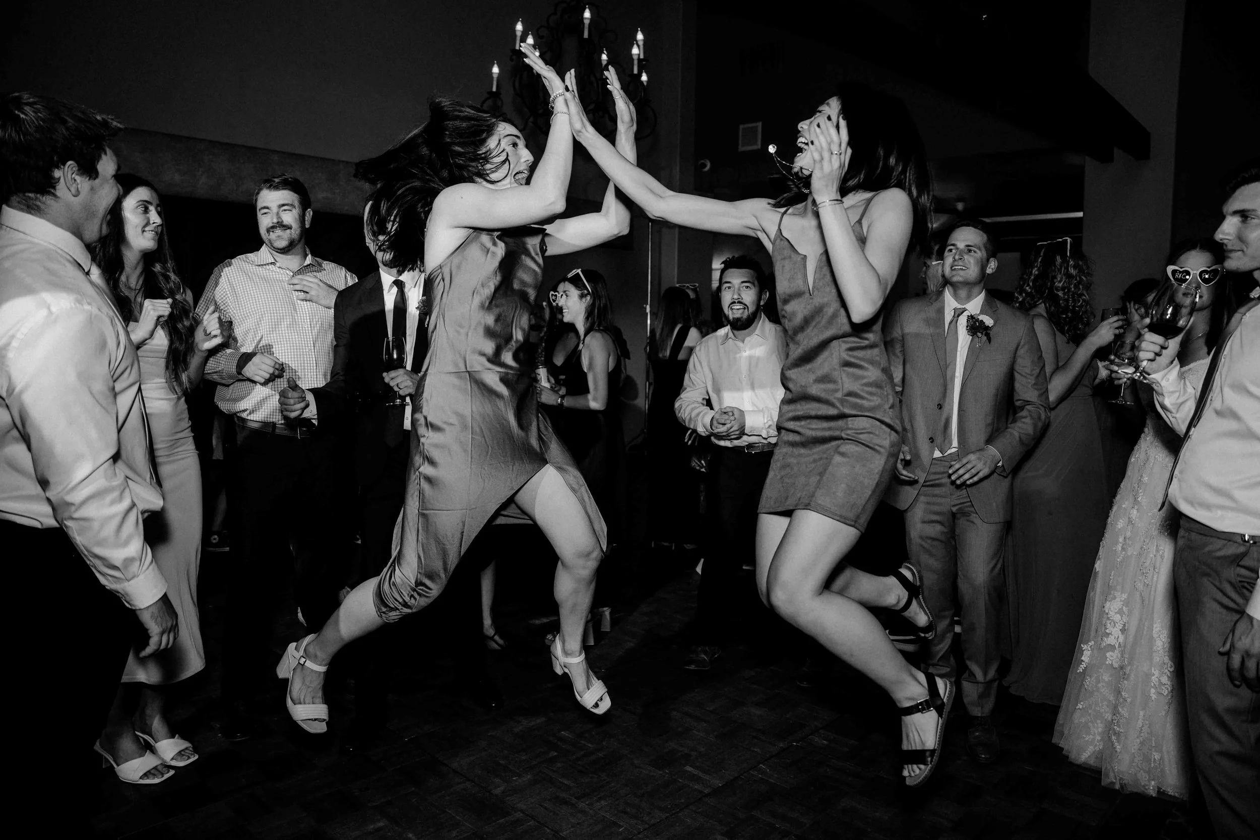 Two women dancing and high-fiving at a party or celebration, surrounded by other guests in formal attire.