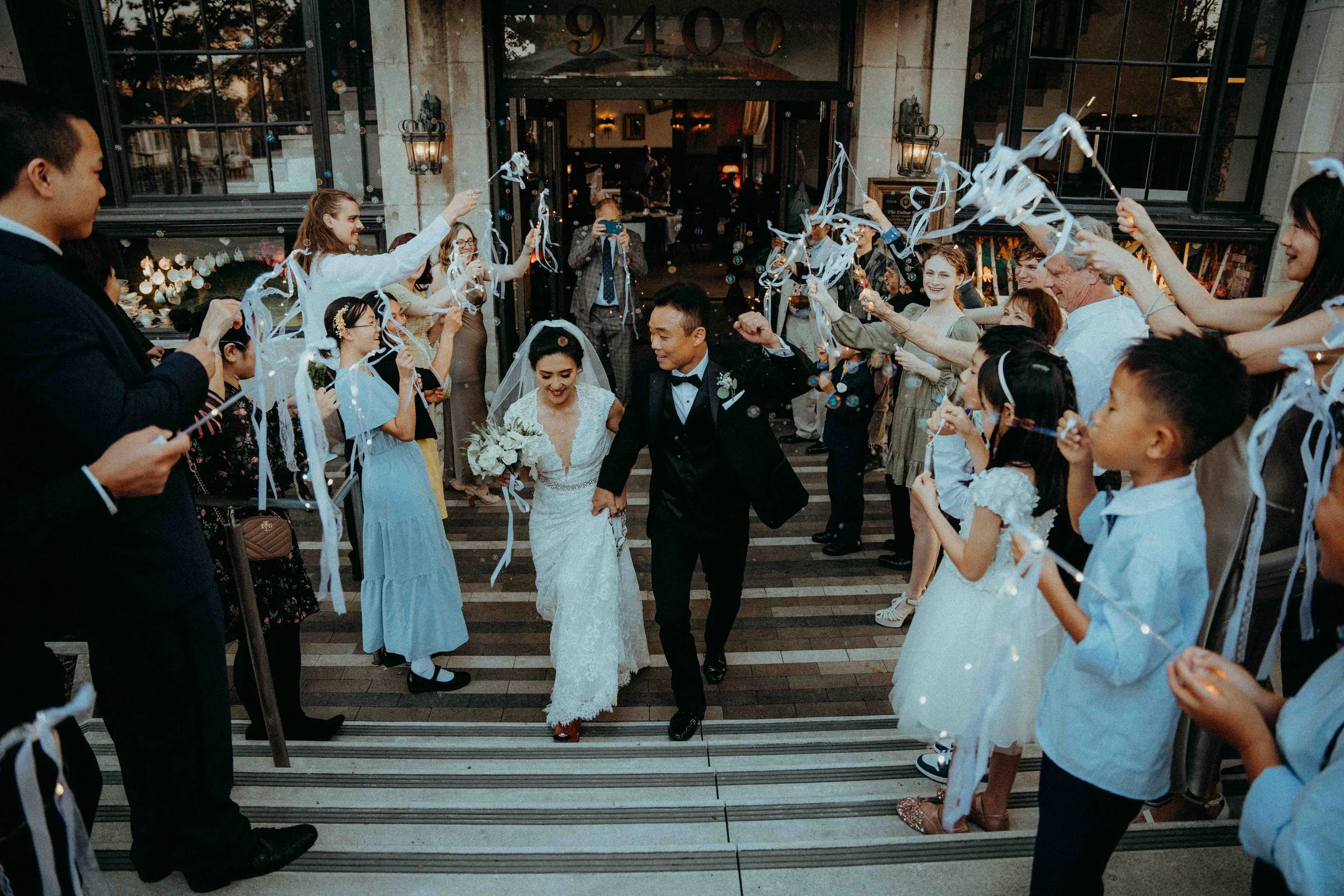 Bride and groom walking through a crowd of people holding streamers and bubbles outside a building, celebrating their wedding.