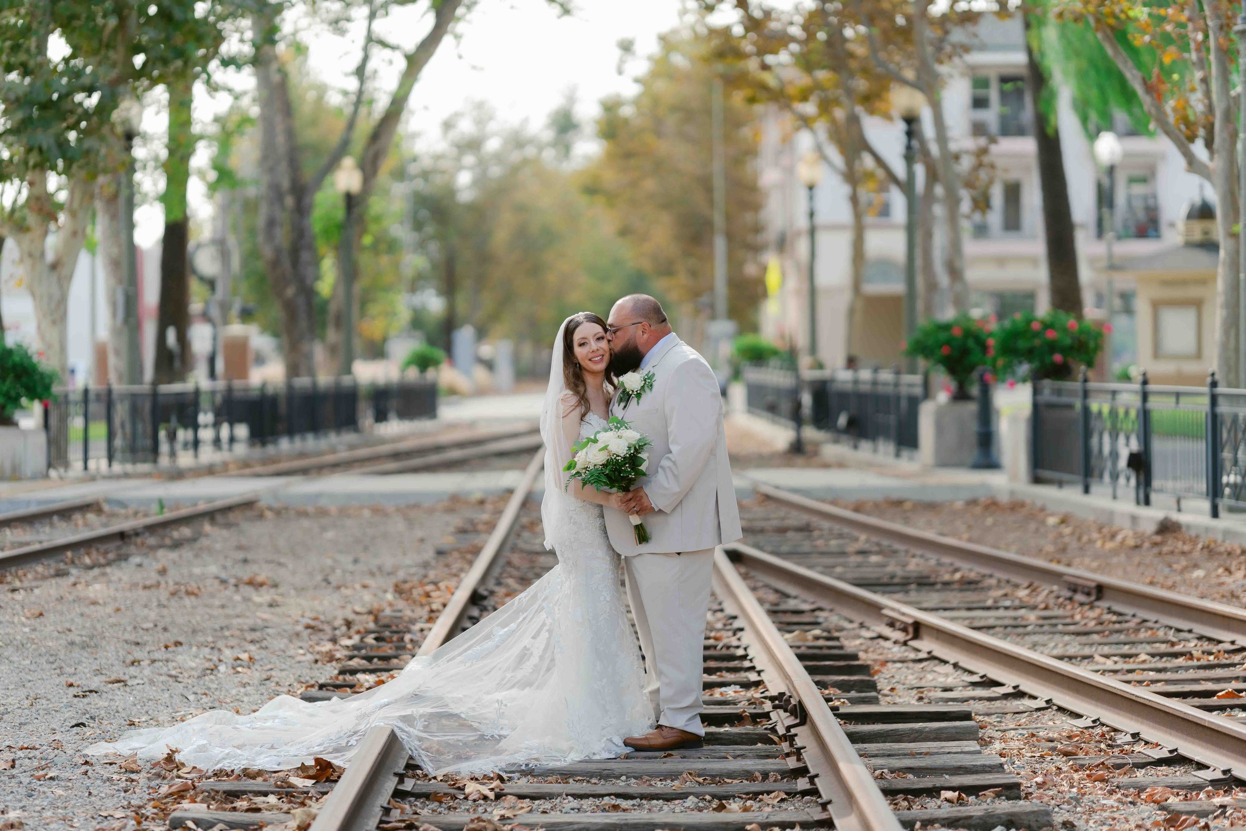 A bride and groom in wedding attire standing on train tracks, sharing a kiss and holding a bouquet of white flowers, in an urban park setting with trees and buildings in the background.