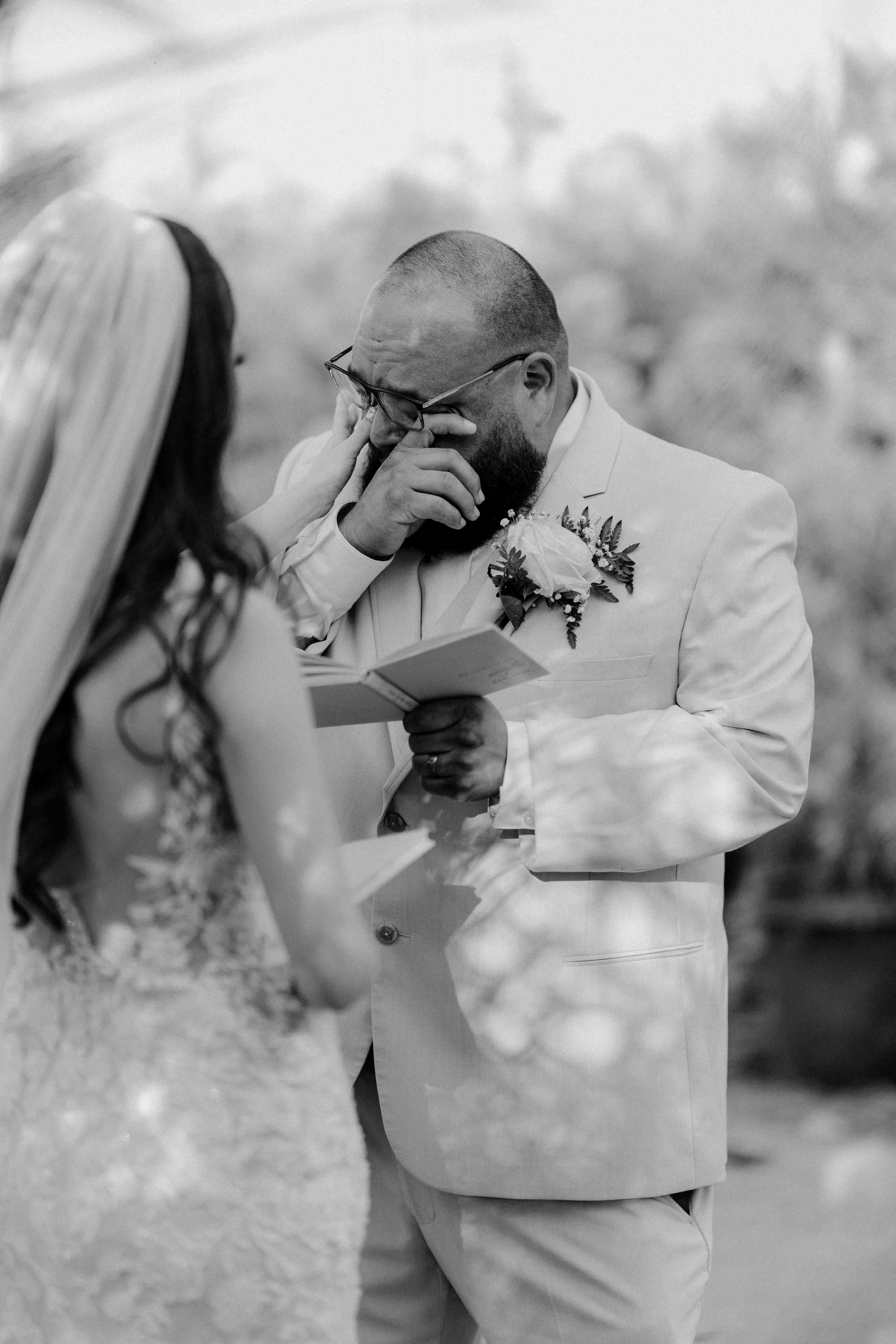 A groom crying during his wedding vows, holding a piece of paper, while the bride faces him, with blurred background.