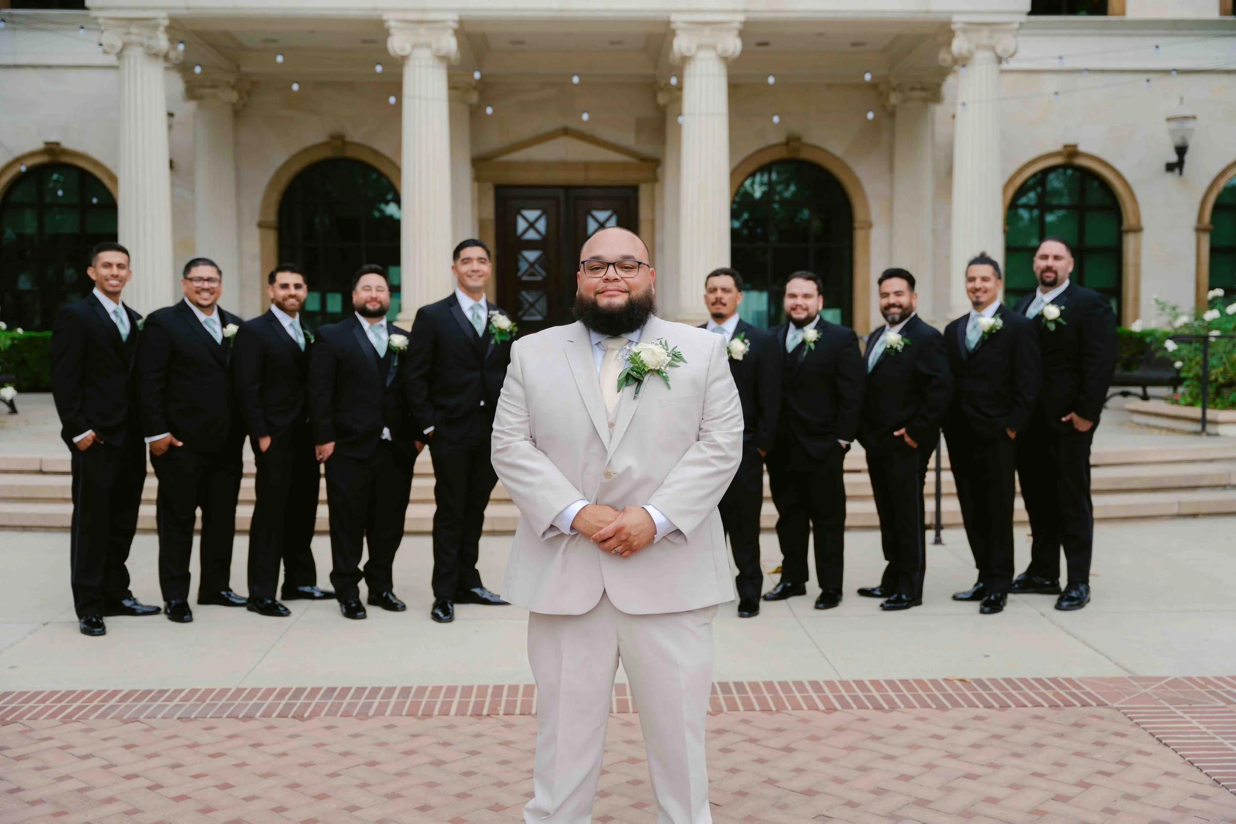 A groom dressed in a white suit stands in front of his wedding party, with groomsmen in black suits and ties, in front of a building with columns and stairs.