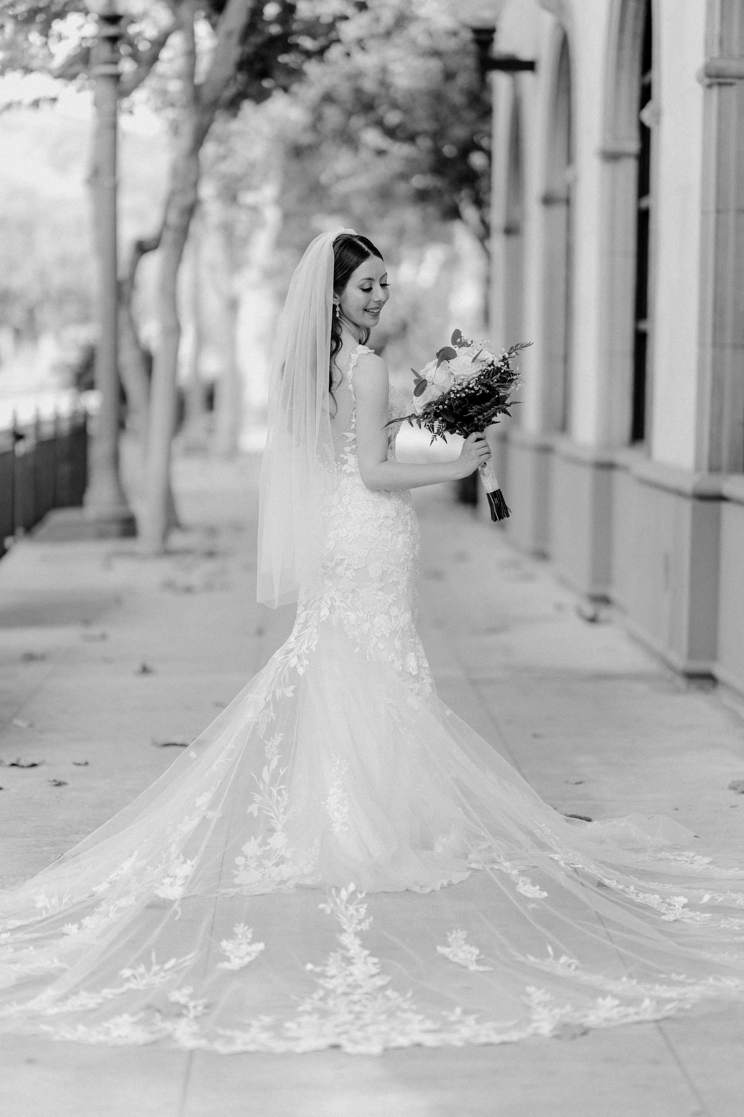 A bride in a lace wedding dress and veil holding a bouquet, standing on a sidewalk with trees and buildings in the background, smiling in black and white.