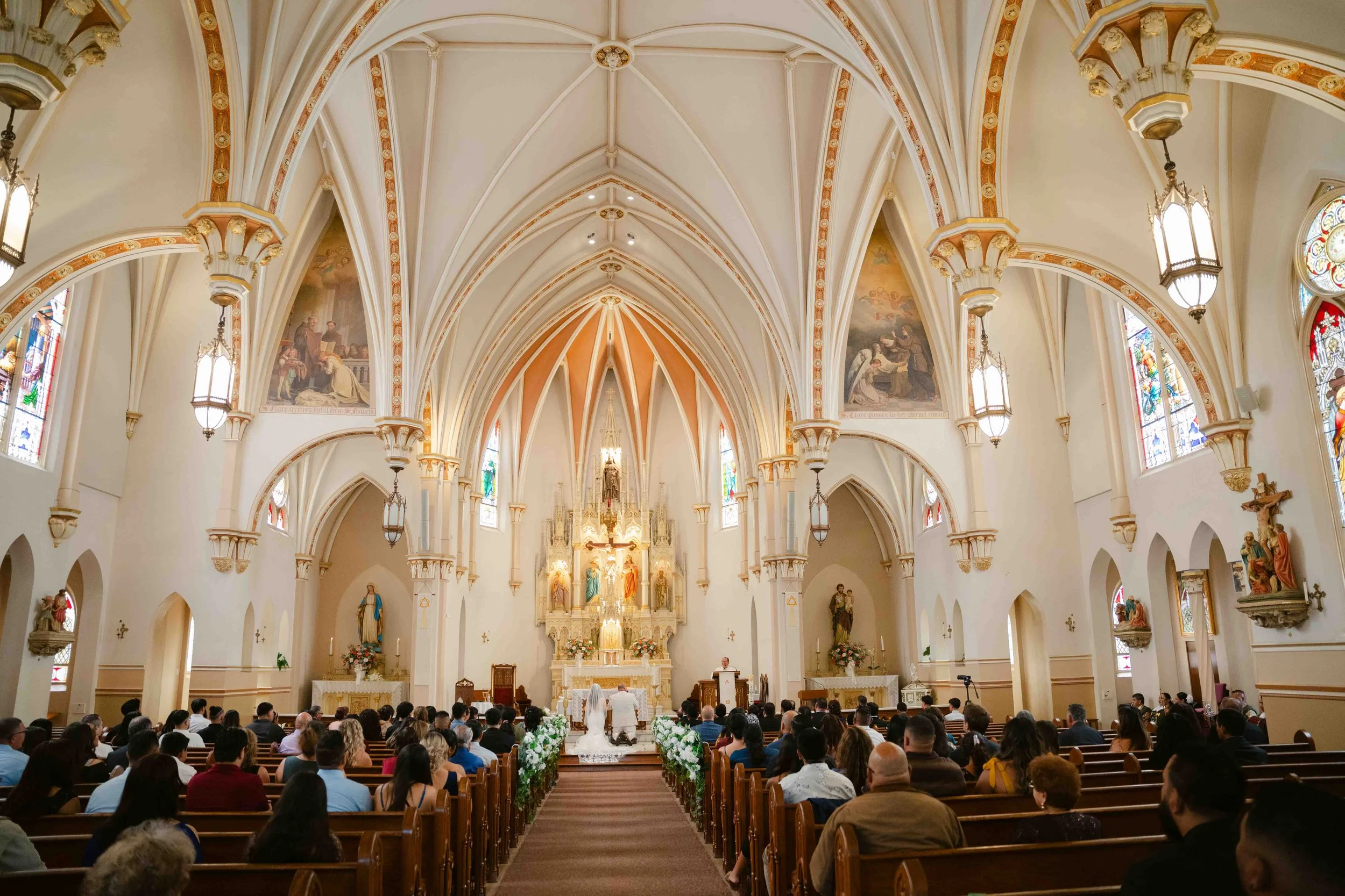 Interior of a church during a wedding ceremony with a congregation seated in pews, an altar with religious statues and decorations, stained glass windows, and Gothic architectural details.