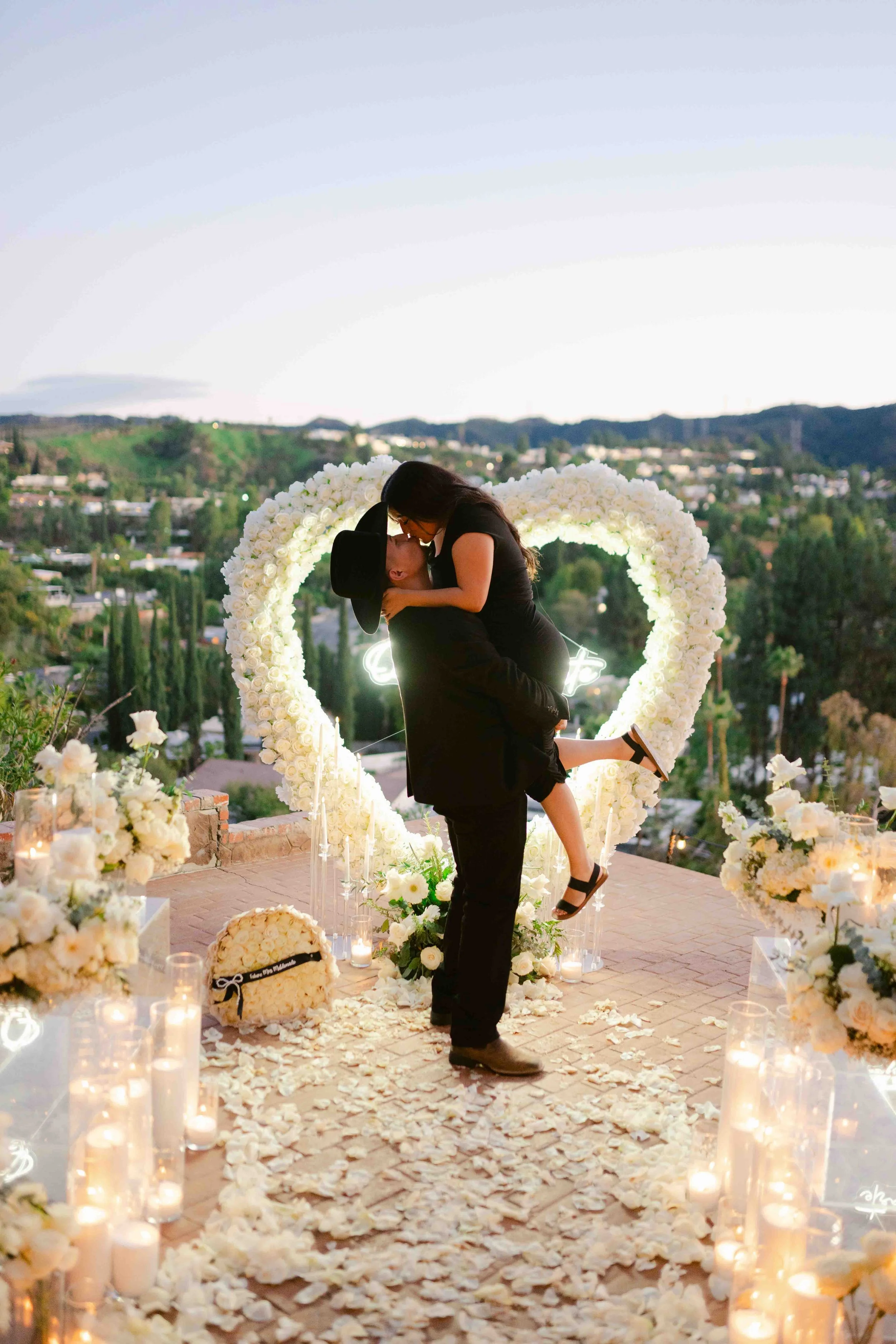 A couple is sharing a romantic kiss at a wedding or engagement celebration, standing in front of a heart-shaped floral arch decorated with white roses, surrounded by candles and flower petals on the ground. The scene takes place outdoors with a sceni