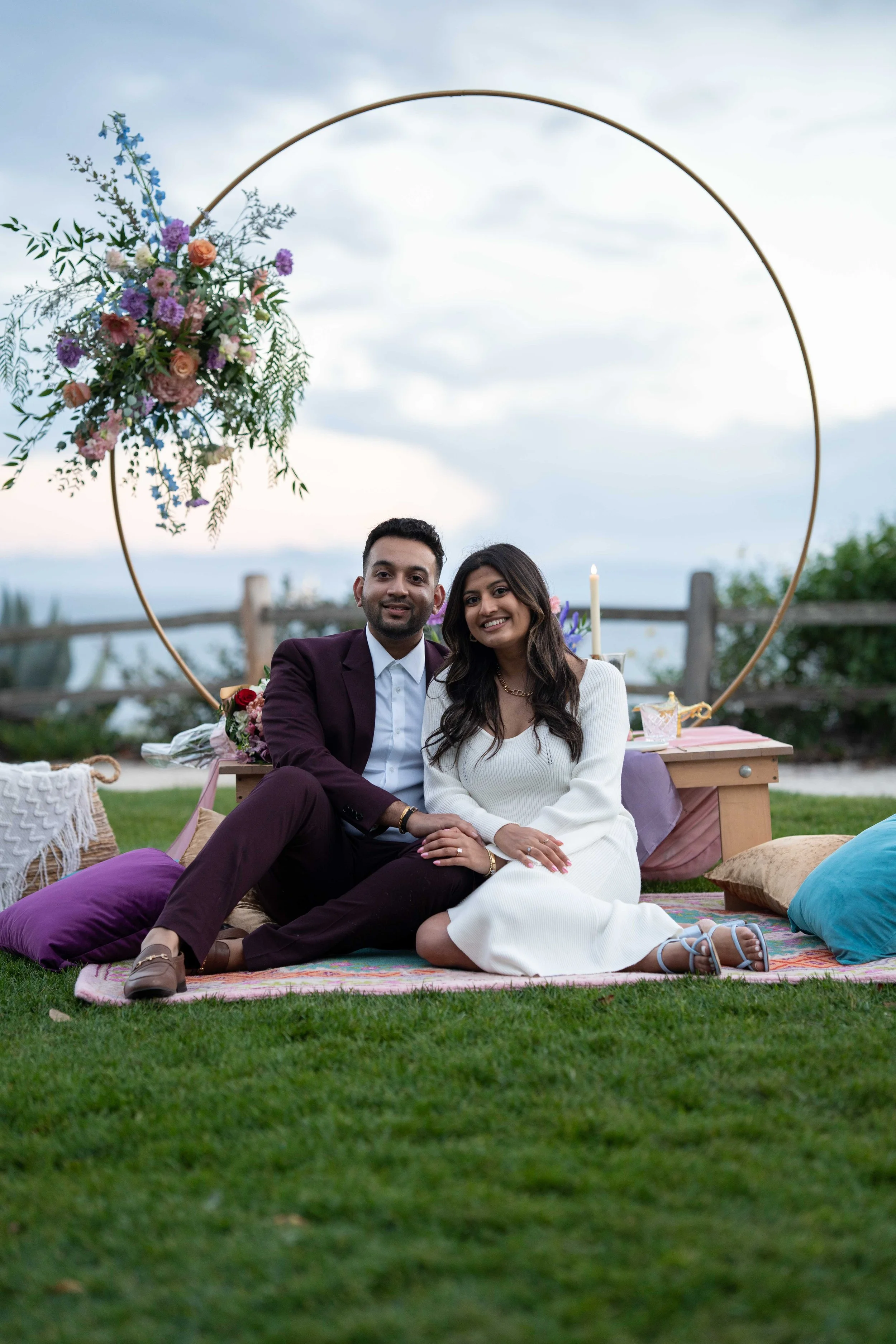 A happy couple sitting on a blanket outdoors at dusk, with a decorative ring structure, flowers, candles, and pillows around them.