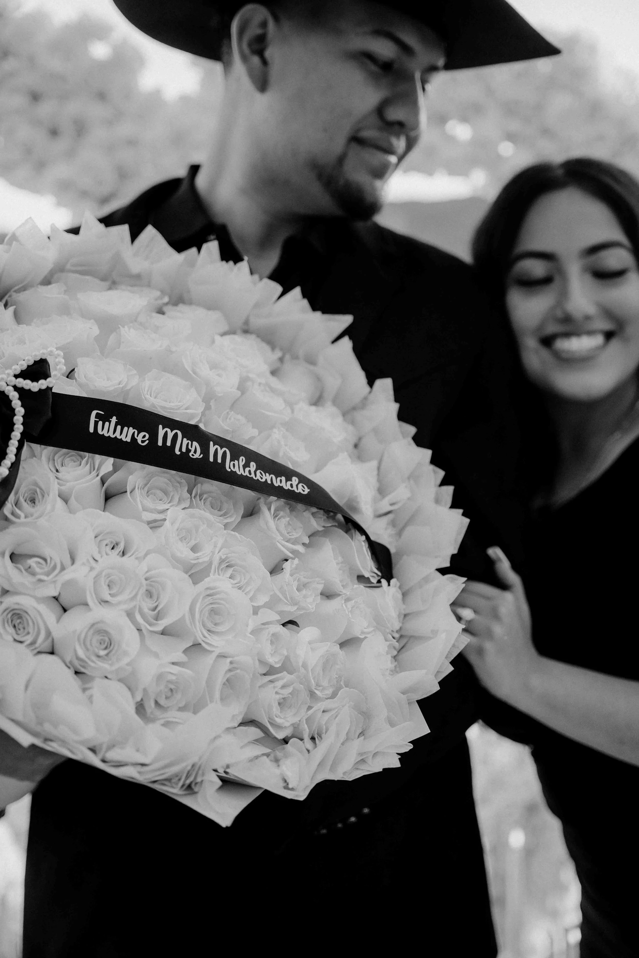 Black and white photo of a smiling man in a cowboy hat holding a bouquet of white roses with a ribbon that reads "Future Mrs. Maldonado," next to a woman with closed eyes and a smiling expression.