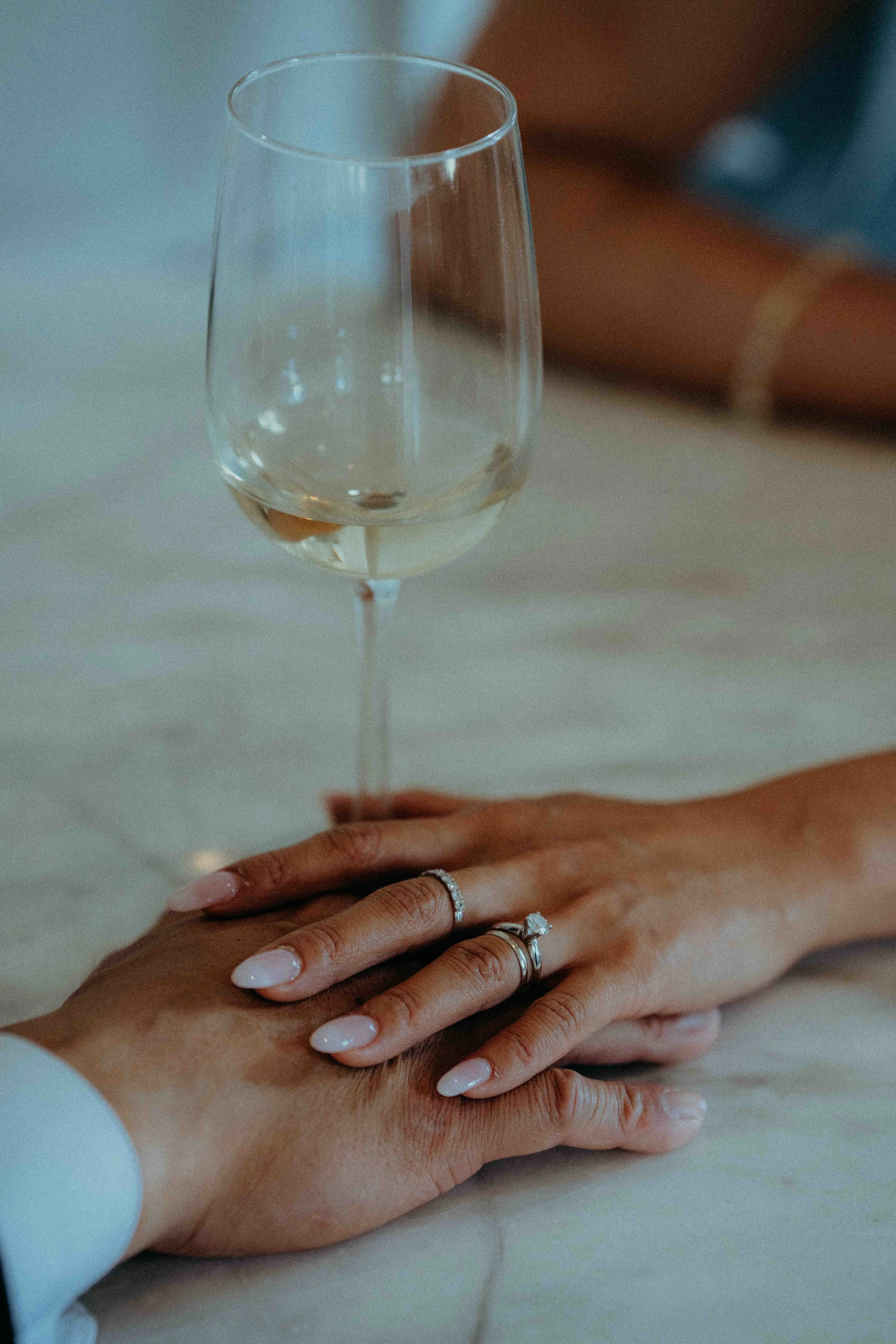 Two hands, one with a diamond engagement ring and a wedding band, resting on a table with a glass of white wine in the background.