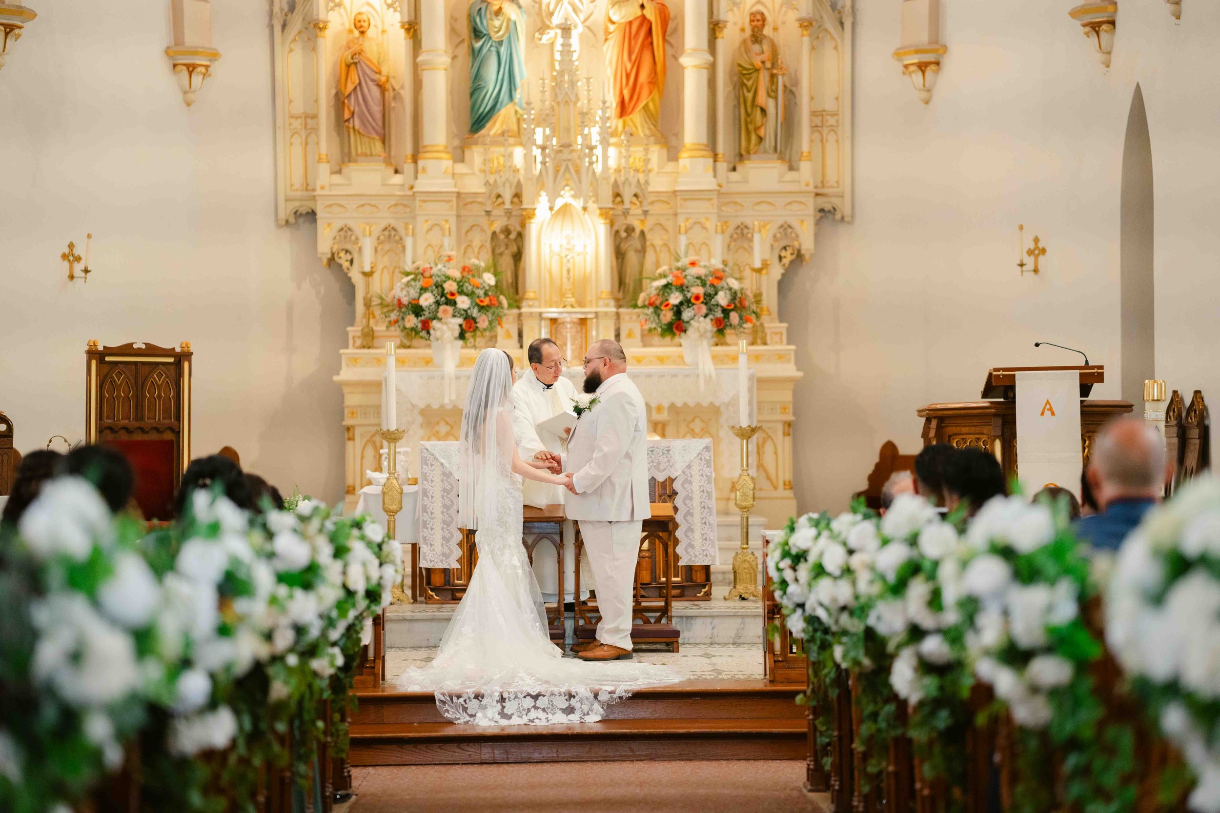 A wedding ceremony taking place in a church with a bride and groom holding hands before an officiant. The church has an ornate altar decorated with flowers, candles, and religious statues. Guests are seated on either side, dressed formally, and the s