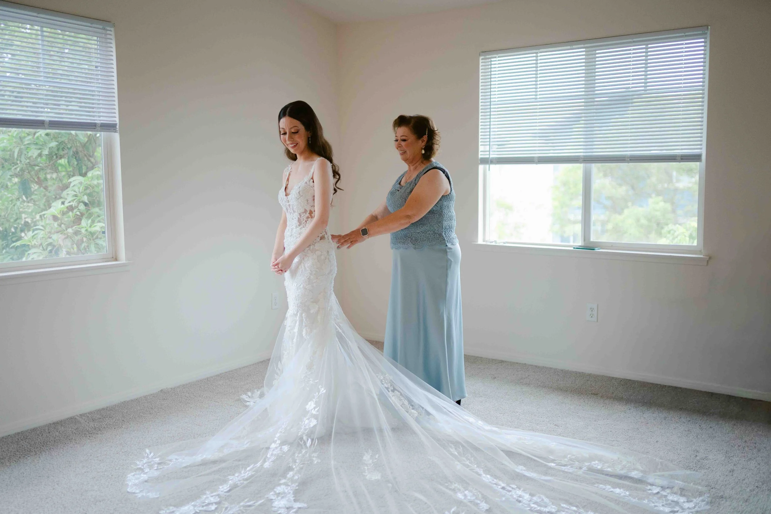 A bride in a white lace wedding dress standing next to an older woman, likely her mother, in a light blue gown, in a bright room with large windows and blinds, as they prepare for the wedding.