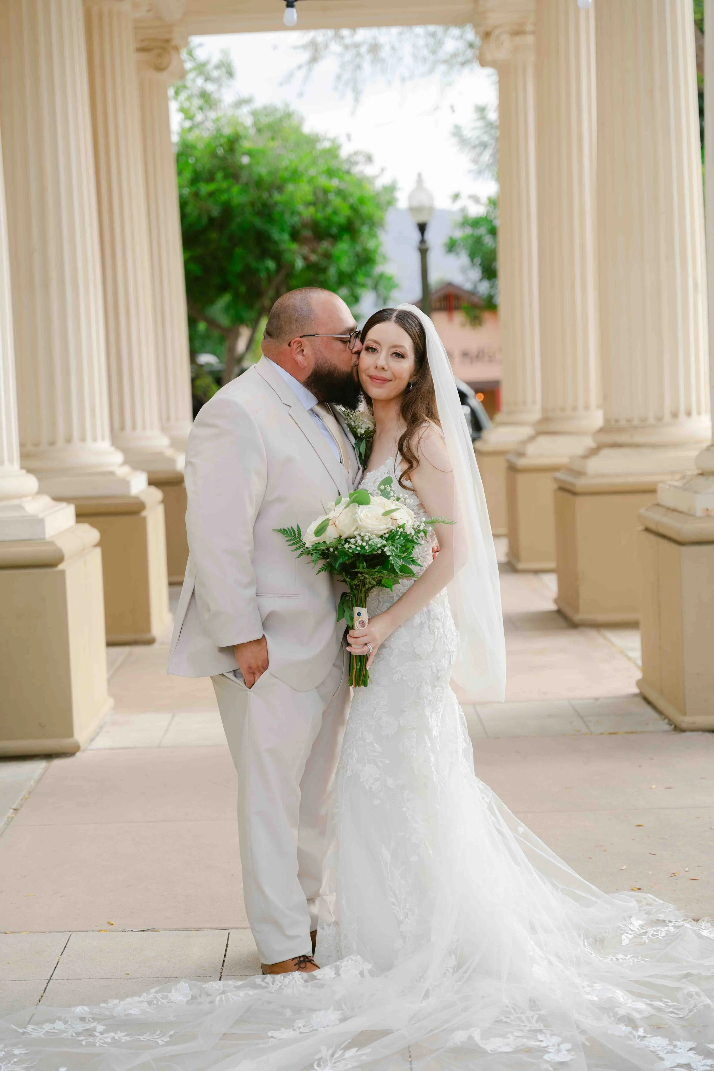 A bride and groom in wedding attire standing under cream-colored columns. The groom is kissing the bride's temple. She is holding a bouquet of white flowers and greenery, and is wearing a lace wedding dress with a long train and a veil.