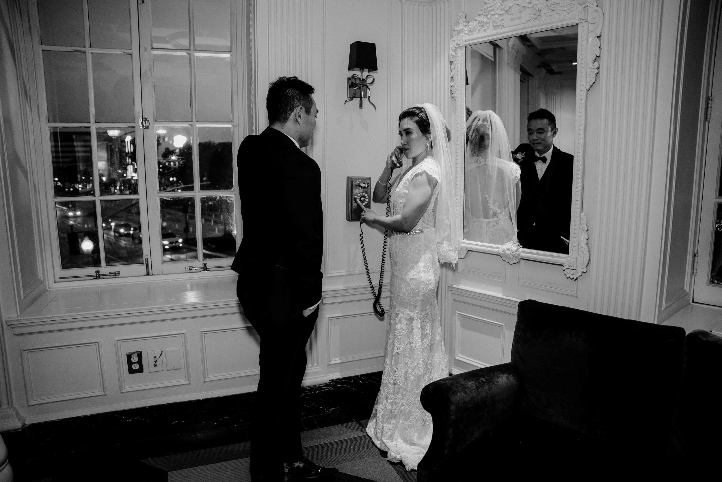 A bride in a lace wedding dress talking on a telephone inside a room with large windows, a mirror, and elegant decor. A groom in a suit is standing nearby, and a reflection of the bride and groom is seen in the mirror.