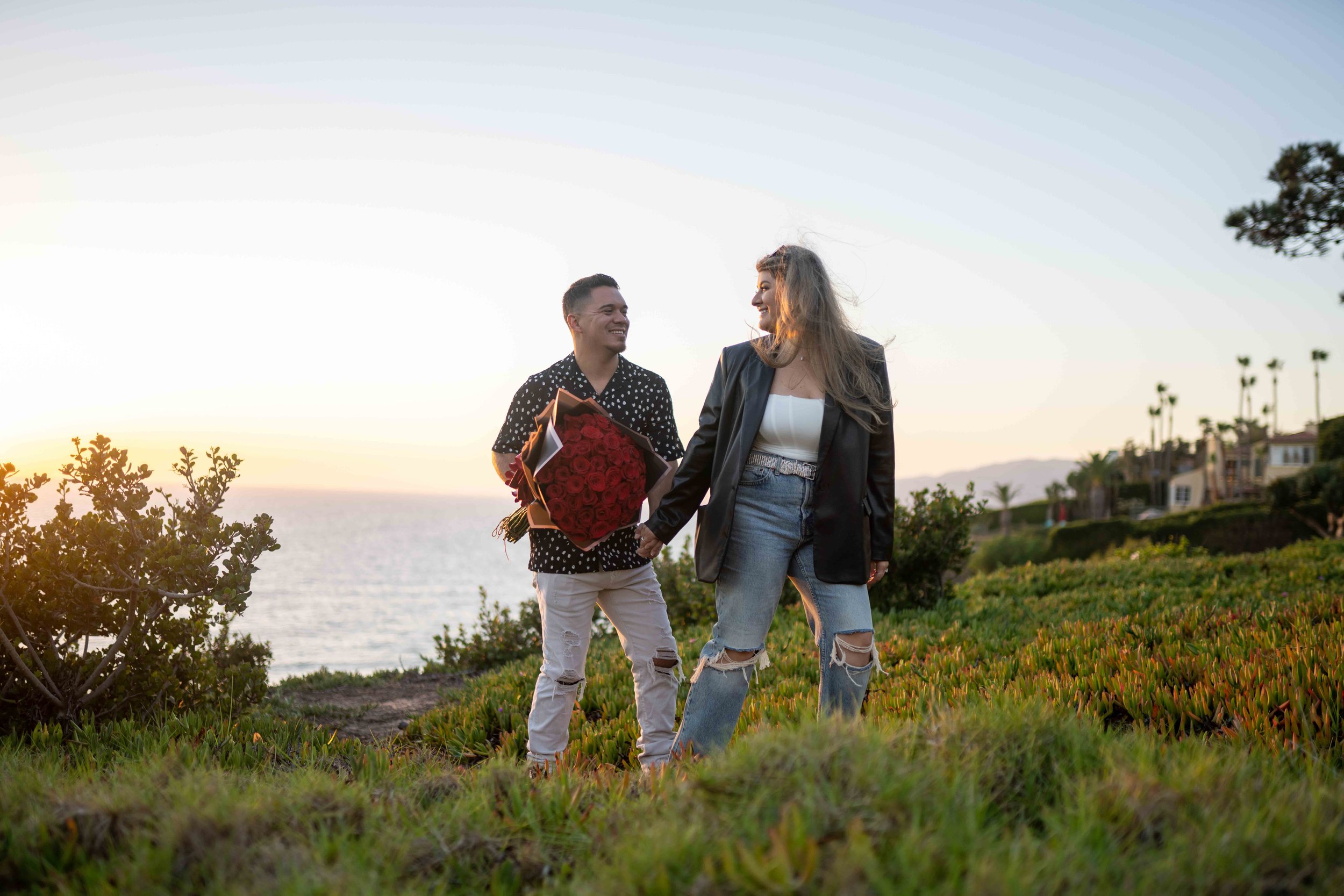 A man holding a large bouquet of red roses and a woman holding hands on a grassy area near the ocean during sunset.