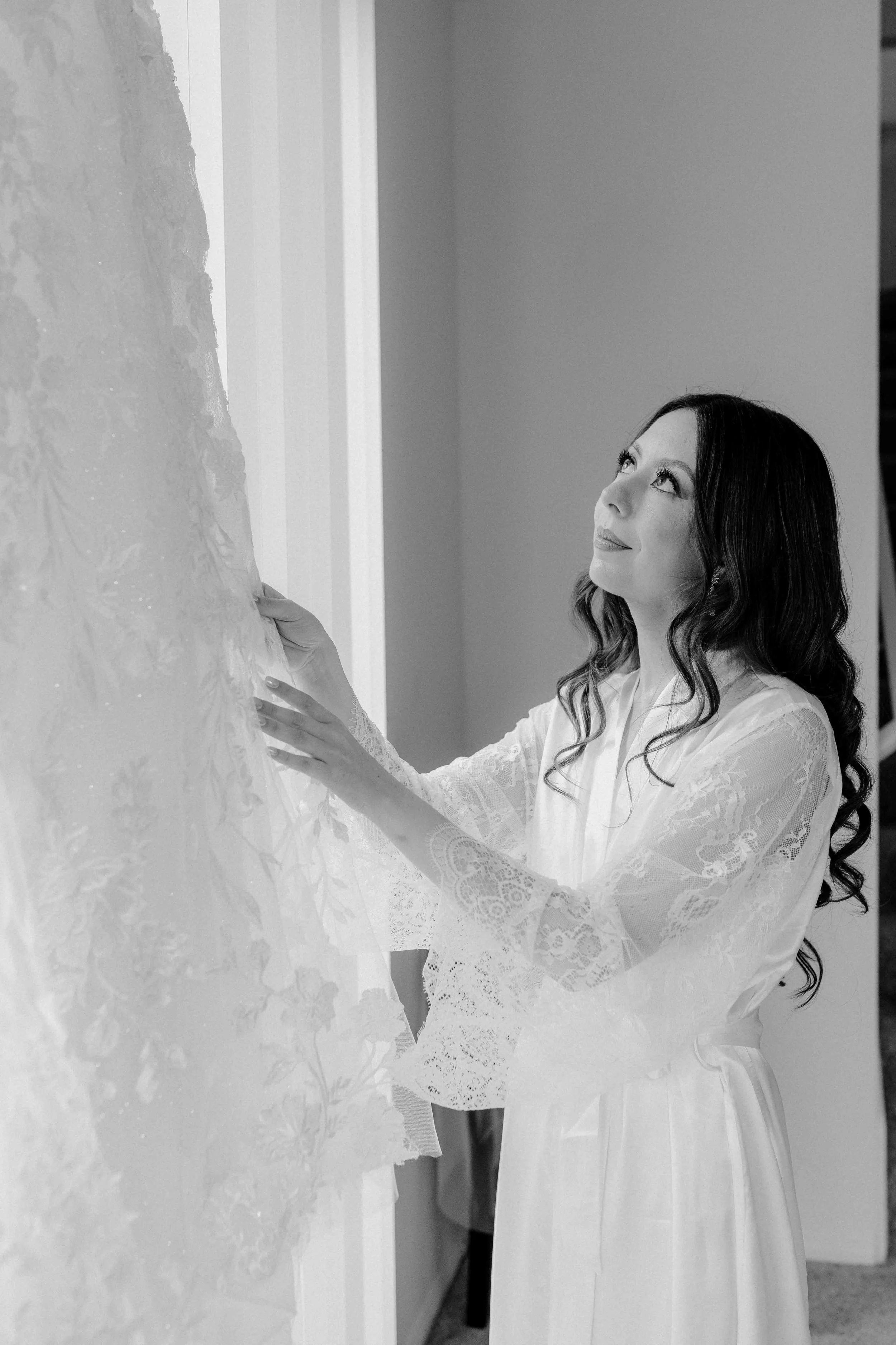 A woman with long dark hair in a satin robe looks at a wedding dress while touching it.