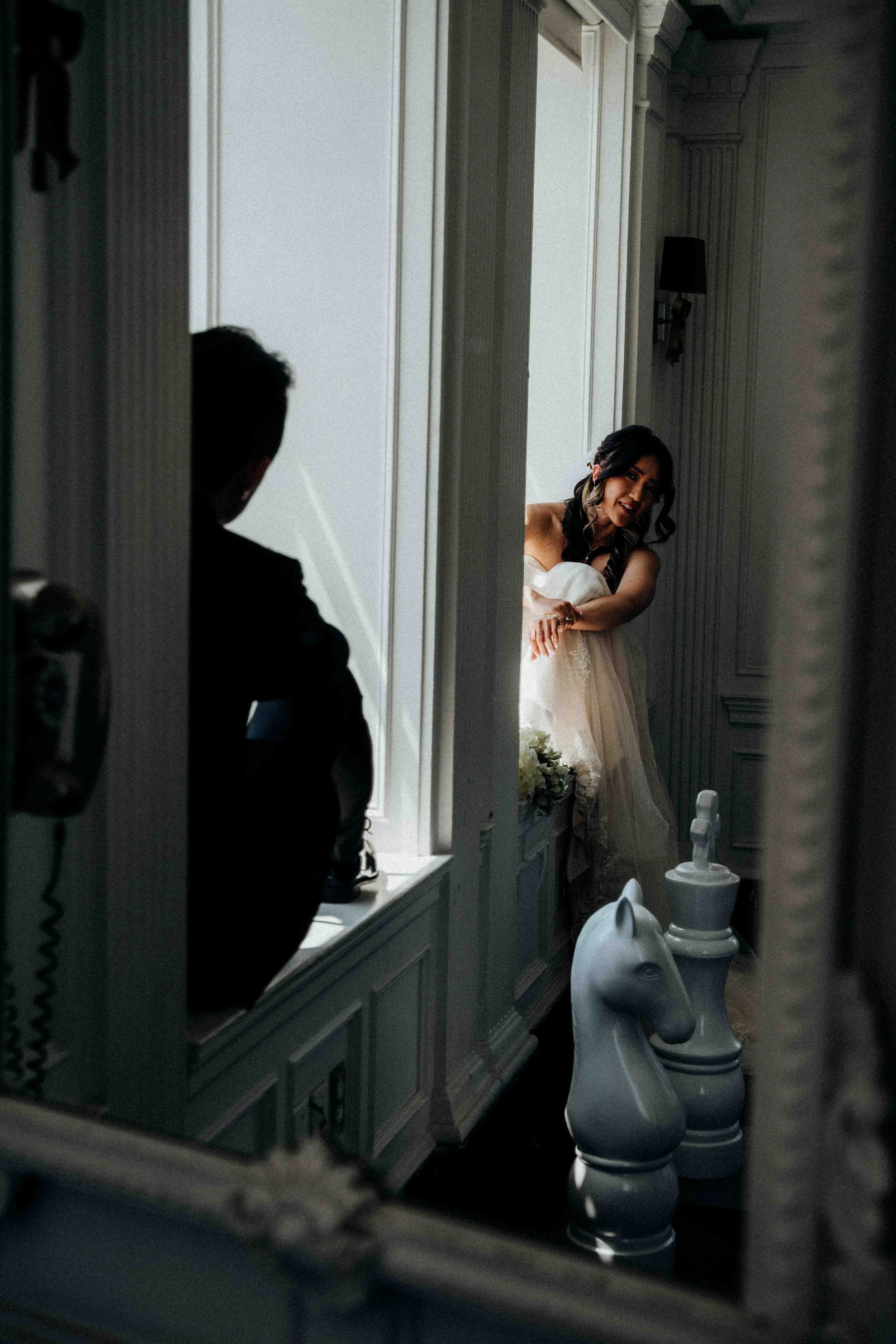 A woman in a wedding dress smiling and leaning on a windowsill, looking at a man sitting nearby in a dark suit, in a room with tall white paneling, a chess piece sculpture, and a horse-shaped object.