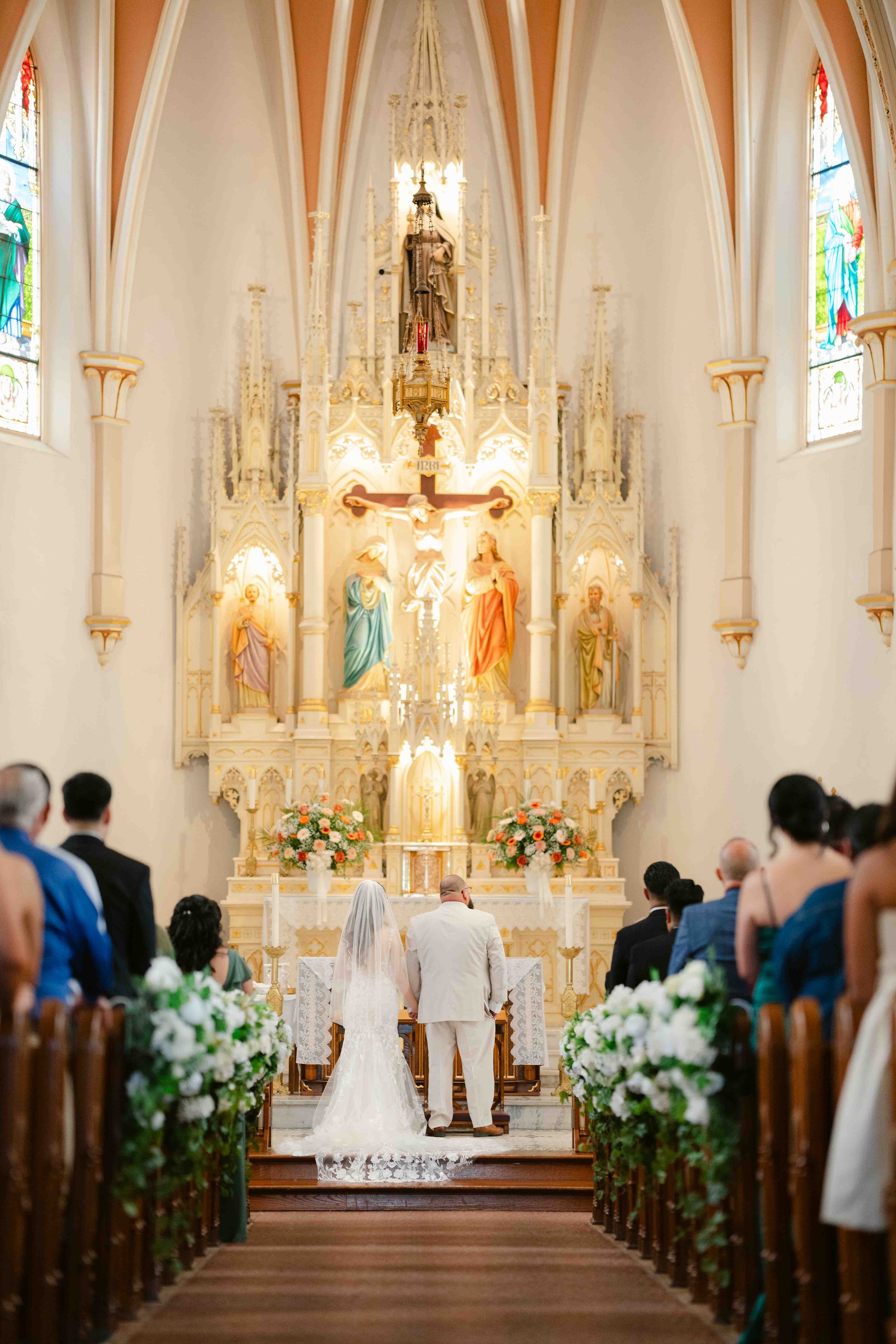 Couple getting married at an altar inside a church, with guests seated on either side, floral arrangements, and ornate religious statues and stained glass windows.