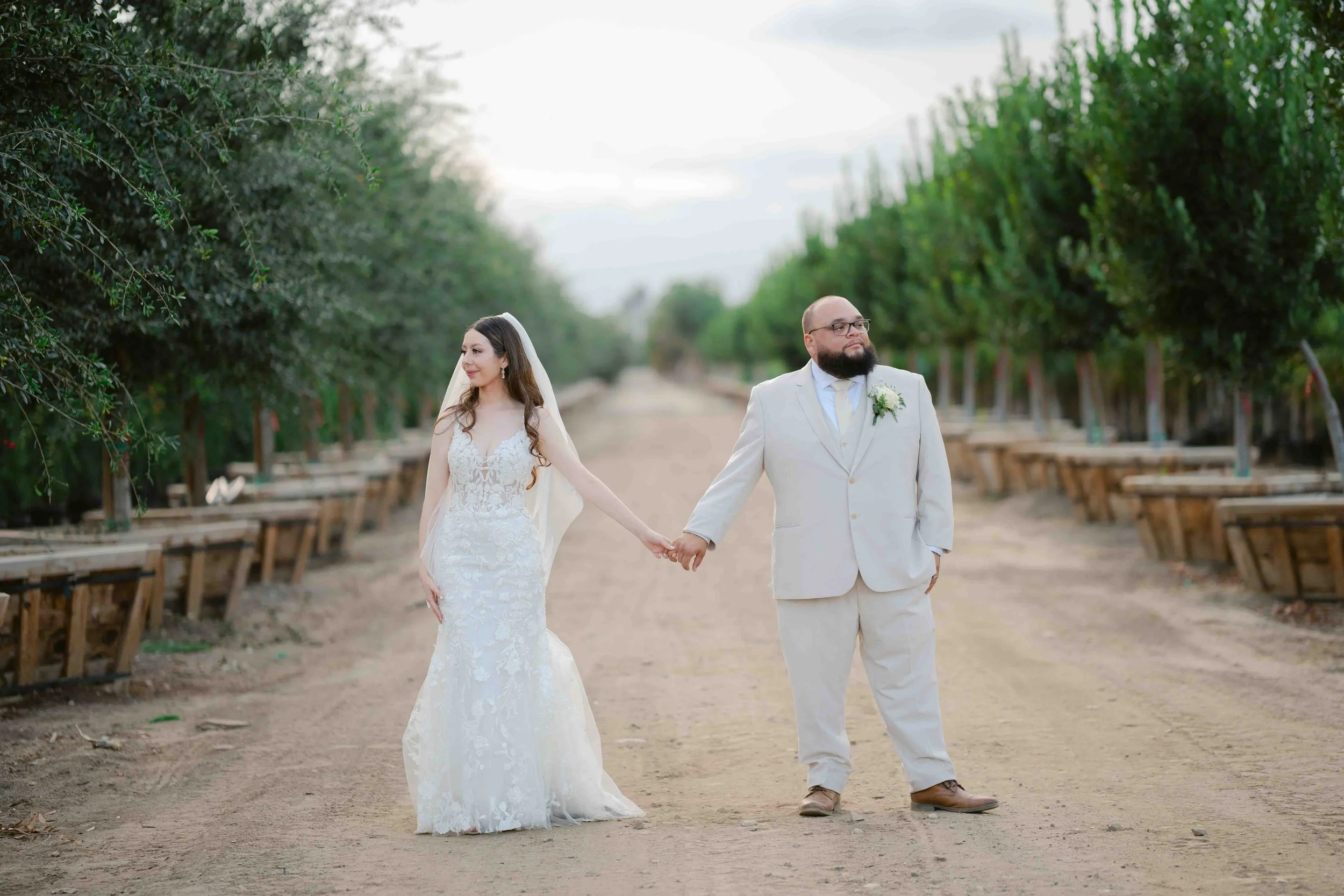 Bride and groom holding hands in a dirt path between rows of trees, outdoor wedding setting.