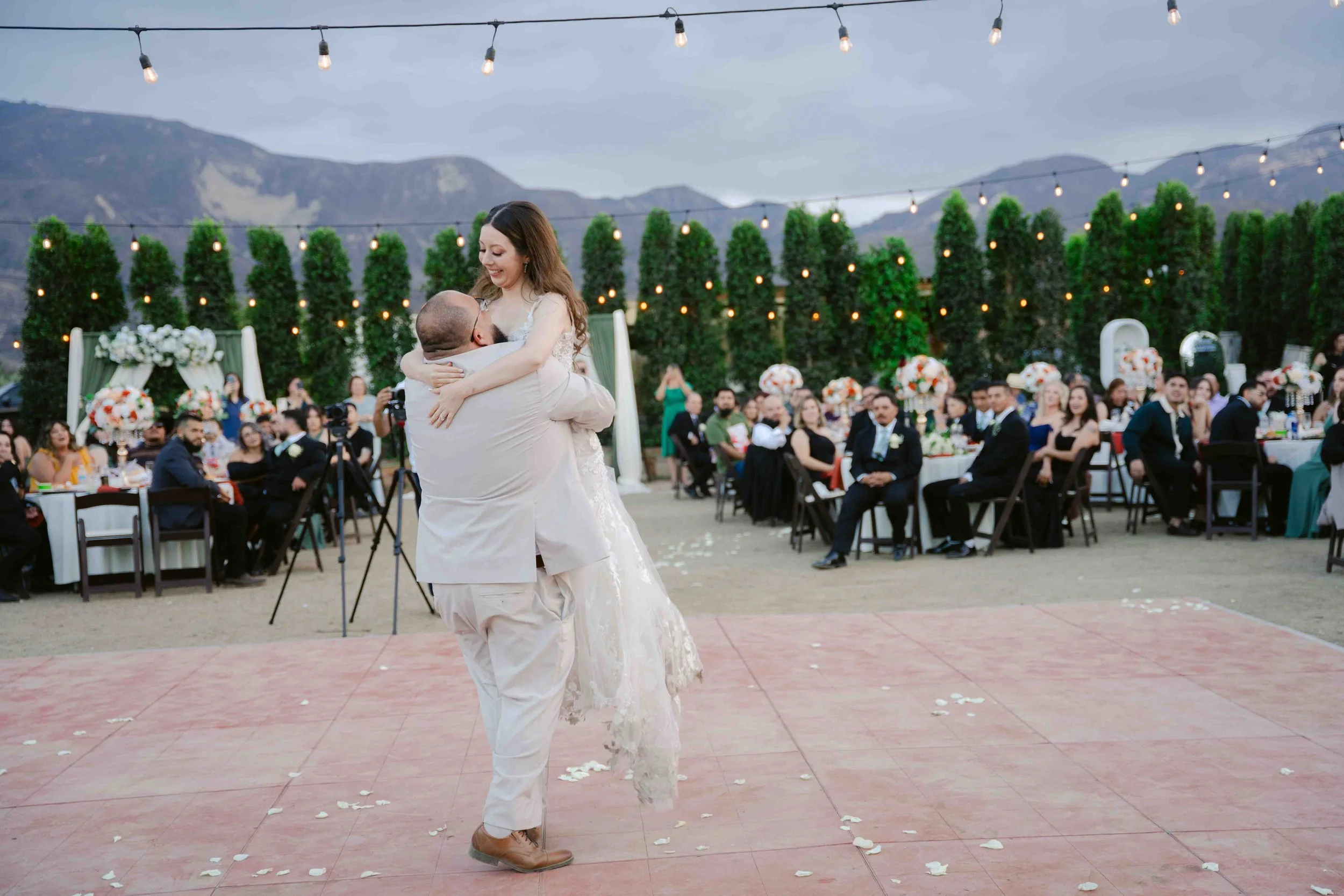 A couple dancing at their wedding reception outdoors, with guests seated at decorated tables, string lights overhead, and mountains in the background.