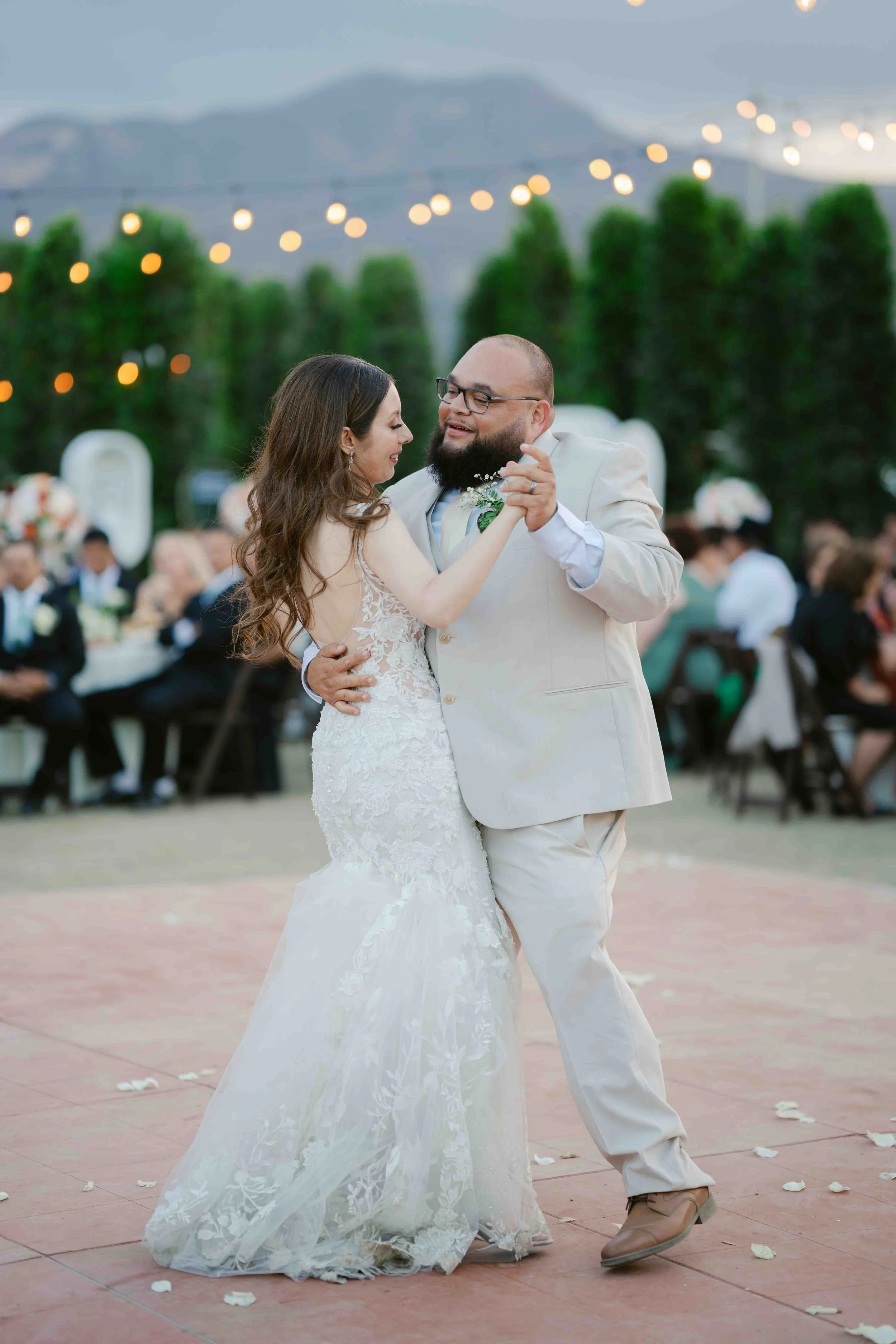 A bride and groom share a dance at their outdoor wedding reception during dusk, with guests seated at tables and string lights overhead.