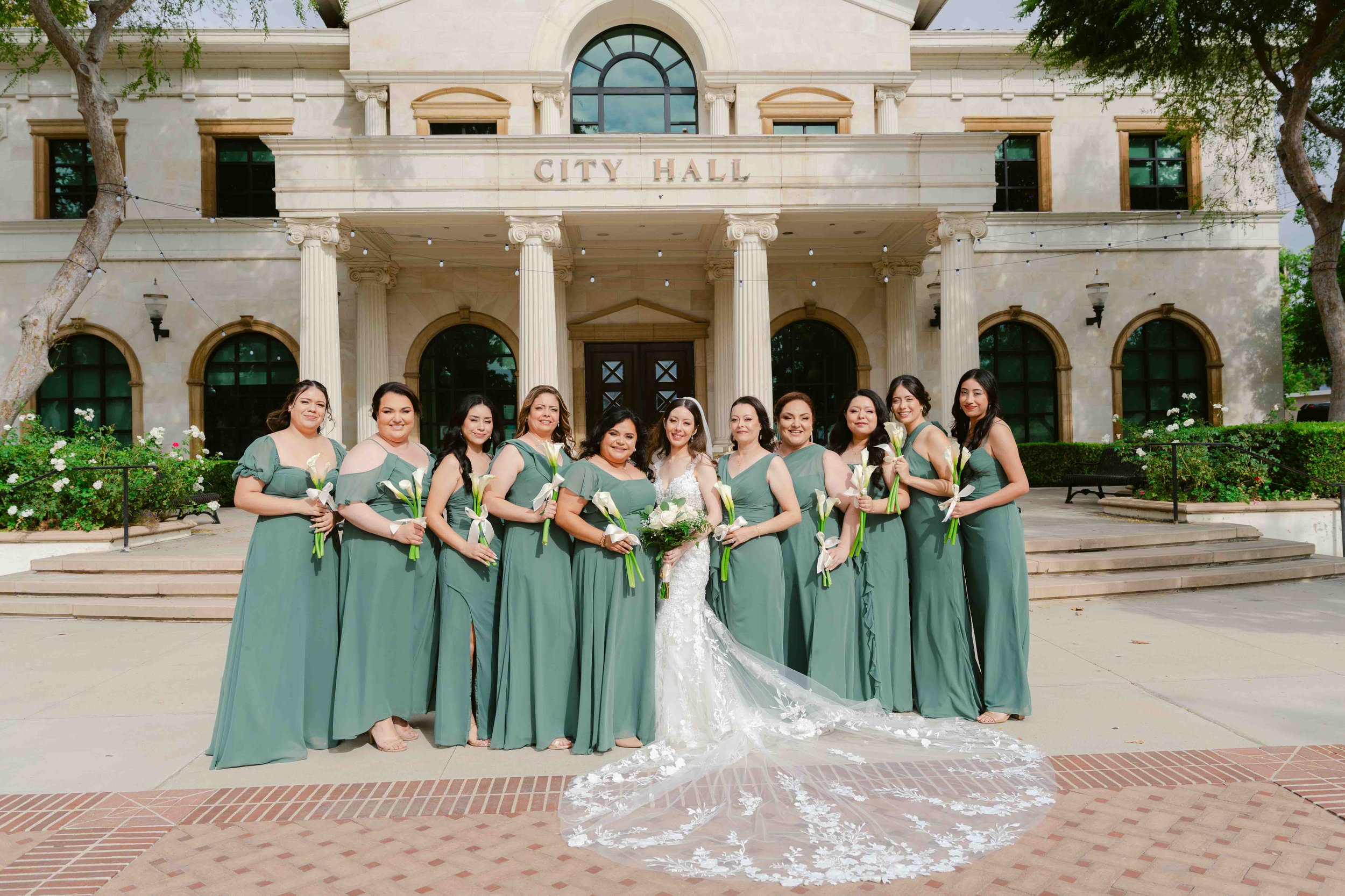 A bride in a white wedding dress and ten bridesmaids in matching teal dresses standing in front of a beige city hall building, holding white calla lily bouquets.