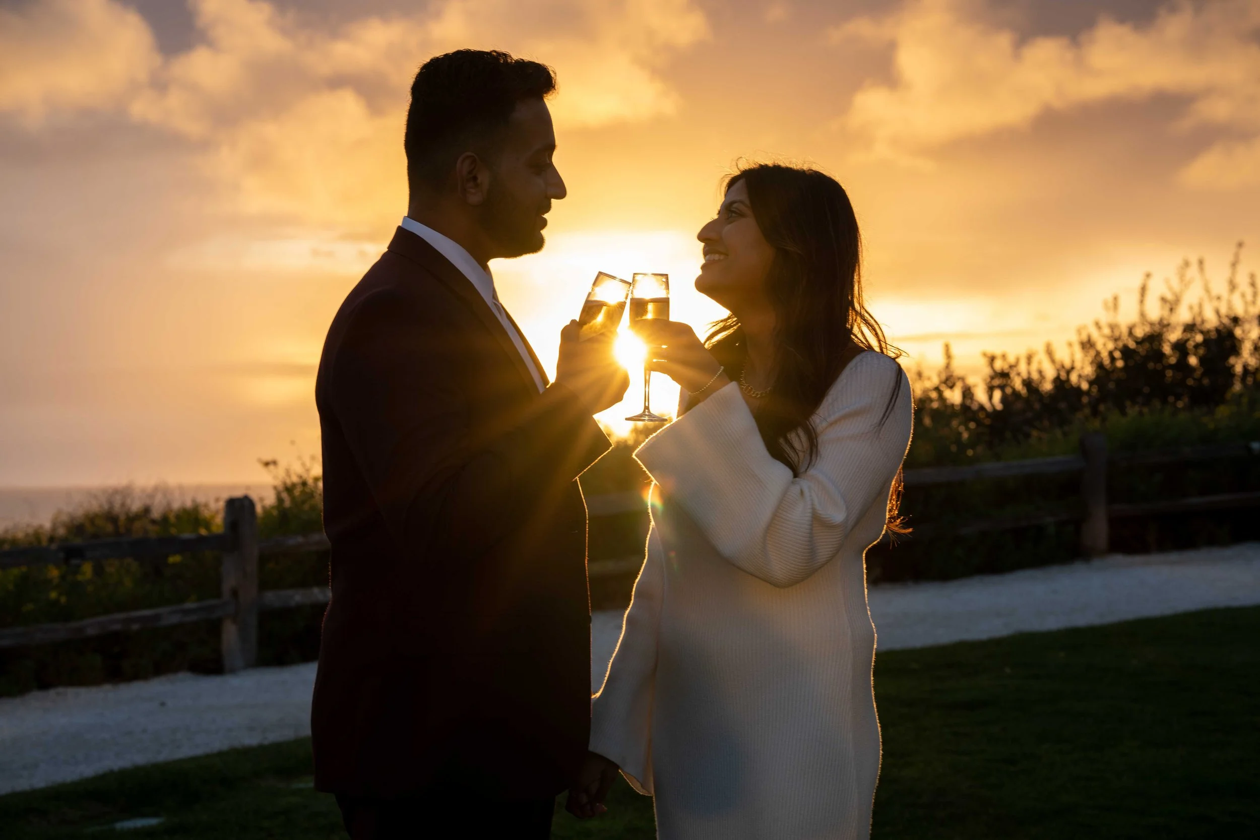 A couple dressed in formal attire toast with champagne glasses during a sunset on a beachside path.