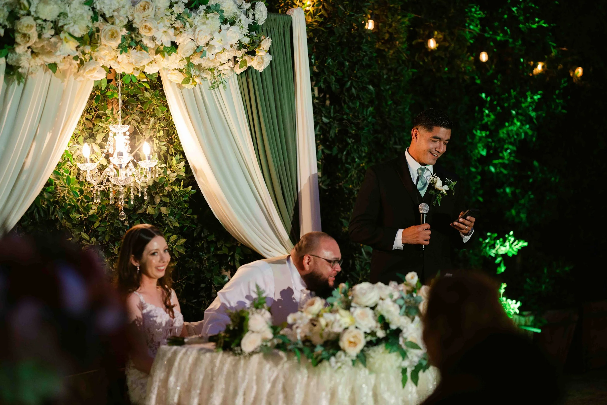 A wedding reception with a bride, groom, and a man giving a speech. The bride has dark hair and is wearing a white dress with lace, smiling. The groom has glasses, a beard, and is wearing a white suit, also smiling. The man giving a speech is wearing
